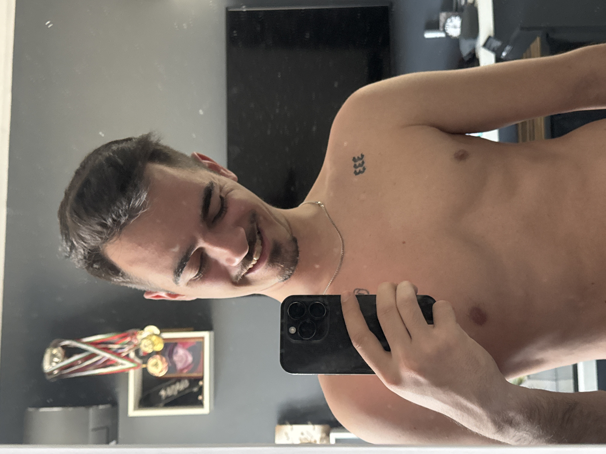 A young adult male is captured in a mirror selfie, shirtless and smiling broadly with his eyes closed, conveying a sense of happiness or amusement. The image is rotated, showing the man's torso and face horizontally across the frame. He has short, dark hair and light stubble on his chin and upper lip. A thin silver chain necklace adorns his neck. A small black tattoo of "333" is visible on his left shoulder/upper arm. He holds a black smartphone, likely an iPhone due to its triple-lens camera module, in his right hand to take the photo.
The setting appears to be an indoor room, possibly a bedroom or hallway, reflected in the mirror. In the background, to the lower left (relative to the man's head), a collection of medals with colorful ribbons (red, white, green, blue) and gold-colored medallions is prominently displayed. Next to the medals, a framed object is partially visible. Further back, a clock with a white face and dark hands is mounted on a wall. Other indistinct household items and a dark rectangular object (possibly a television or large screen) are also reflected.
The image captures a casual, joyful moment. No specific time of day or weather is discernible. The general location is Sofia, Bulgaria.