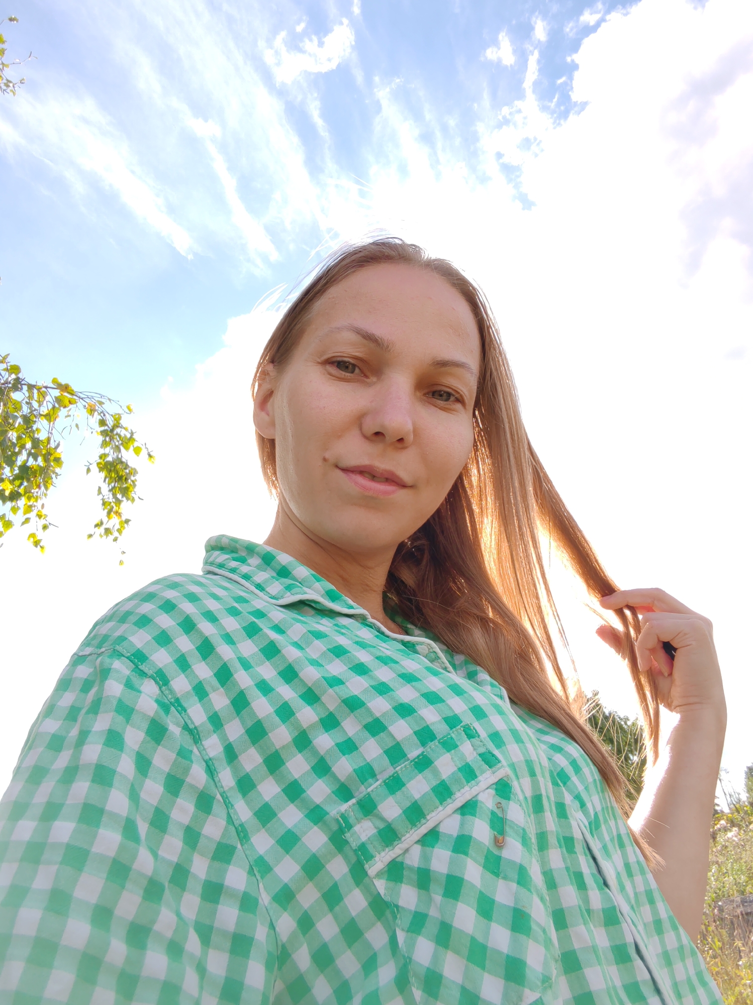 A woman is taking a selfie outdoors in Чухлэм, Russia. She is looking at the camera with a slight smile. She is wearing a green and white checkered shirt. Her right hand is touching her hair, which is lit by the sun. The background shows a bright sky with wispy clouds and some foliage on the left side of the frame. The overall lighting suggests it is daytime, likely late afternoon due to the warm glow on her hair.