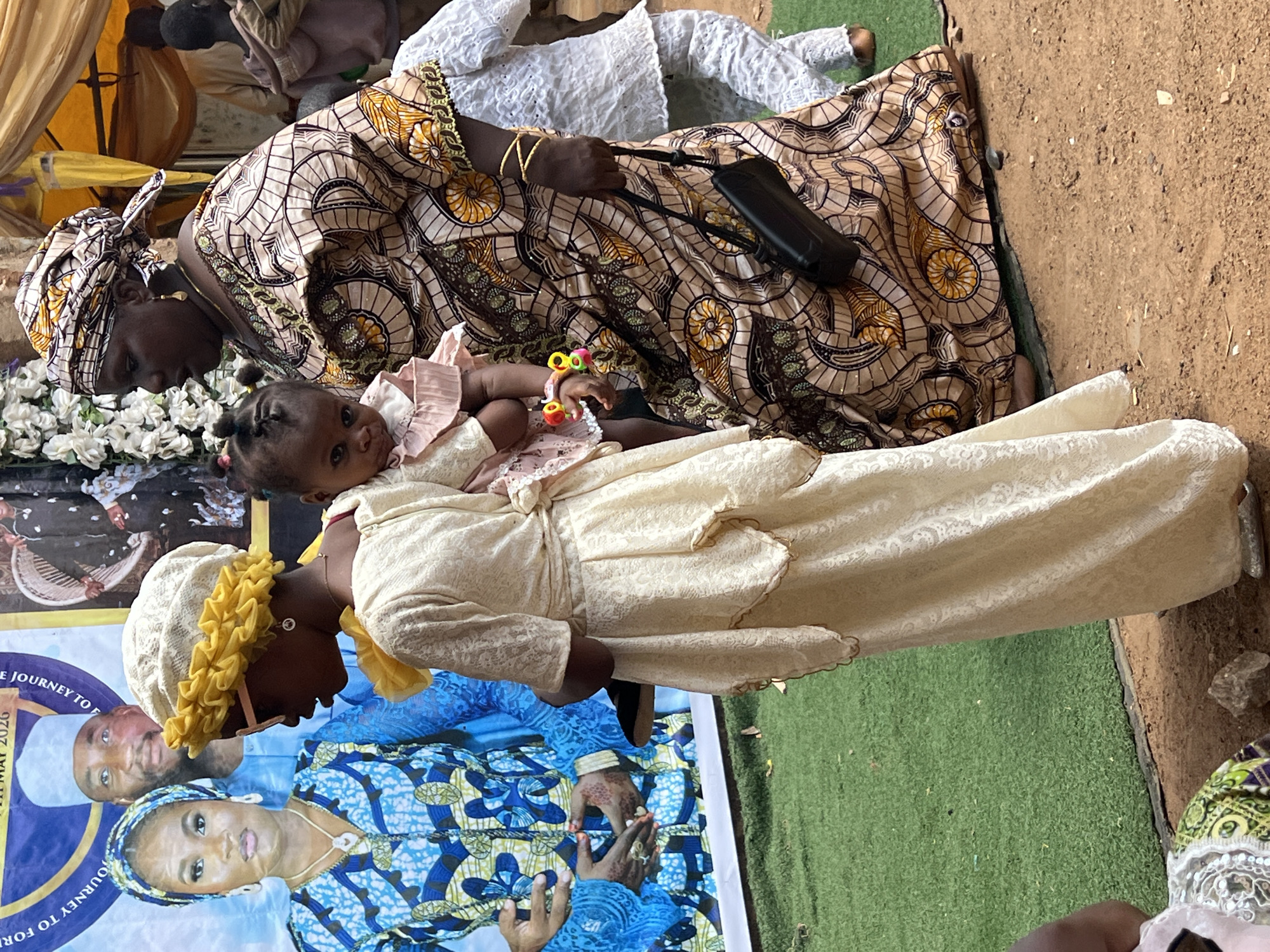A vertical, medium shot captures a scene in Rigasa, Nigeria. Two adult women and a baby are present. One woman, dressed in a cream-colored, full-length dress with a matching head covering adorned with yellow trim, holds a baby wearing a pink dress. The baby looks towards the camera. The other woman, positioned behind the woman and child, is attired in a brown and yellow patterned dress and a matching headwrap. She carries a black purse on her shoulder.
The scene is set on a patch of green artificial turf, bordered by bare earth. A large banner featuring portraits of a man and a woman in traditional clothing is partially visible. The banner includes the text "JOURNEY TO FOR...", with the date "11 MAY 2026" also visible.