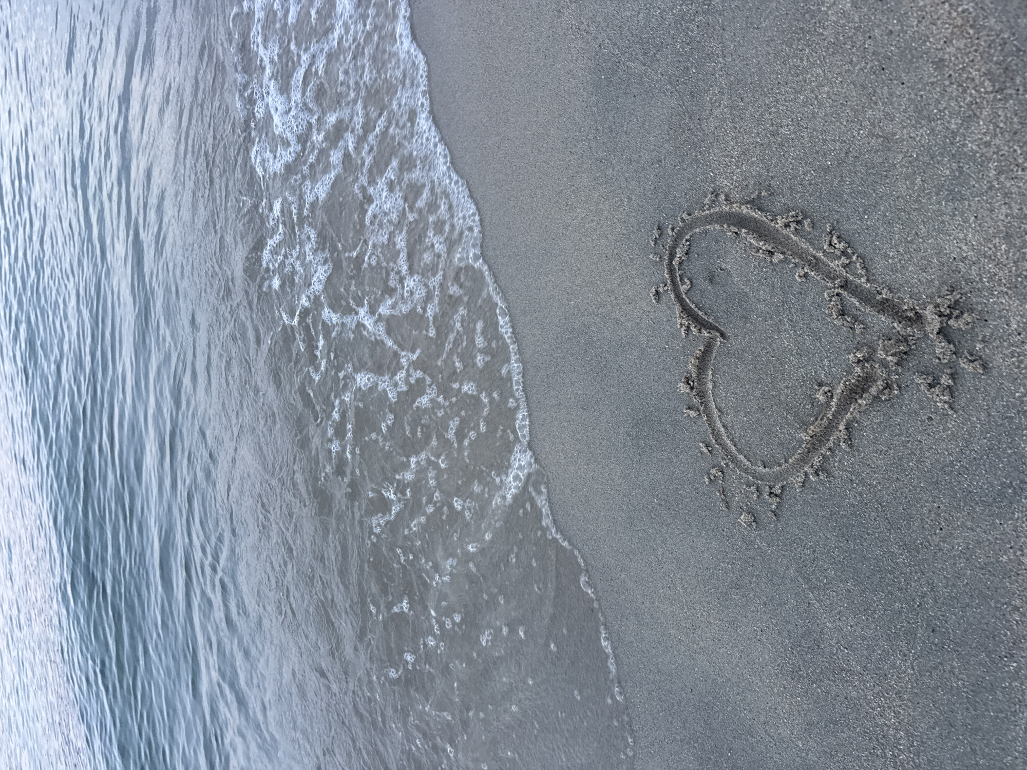 A heart is drawn in the sand on a beach in Keramoti, Greece. The heart is in the lower right portion of the frame, outlined with a slightly darker shade of sand. The sea is in the upper part of the image, with gentle waves lapping at the shore. The water is a muted blue-gray, and the sand is a fine, dark gray. The sky is not visible.