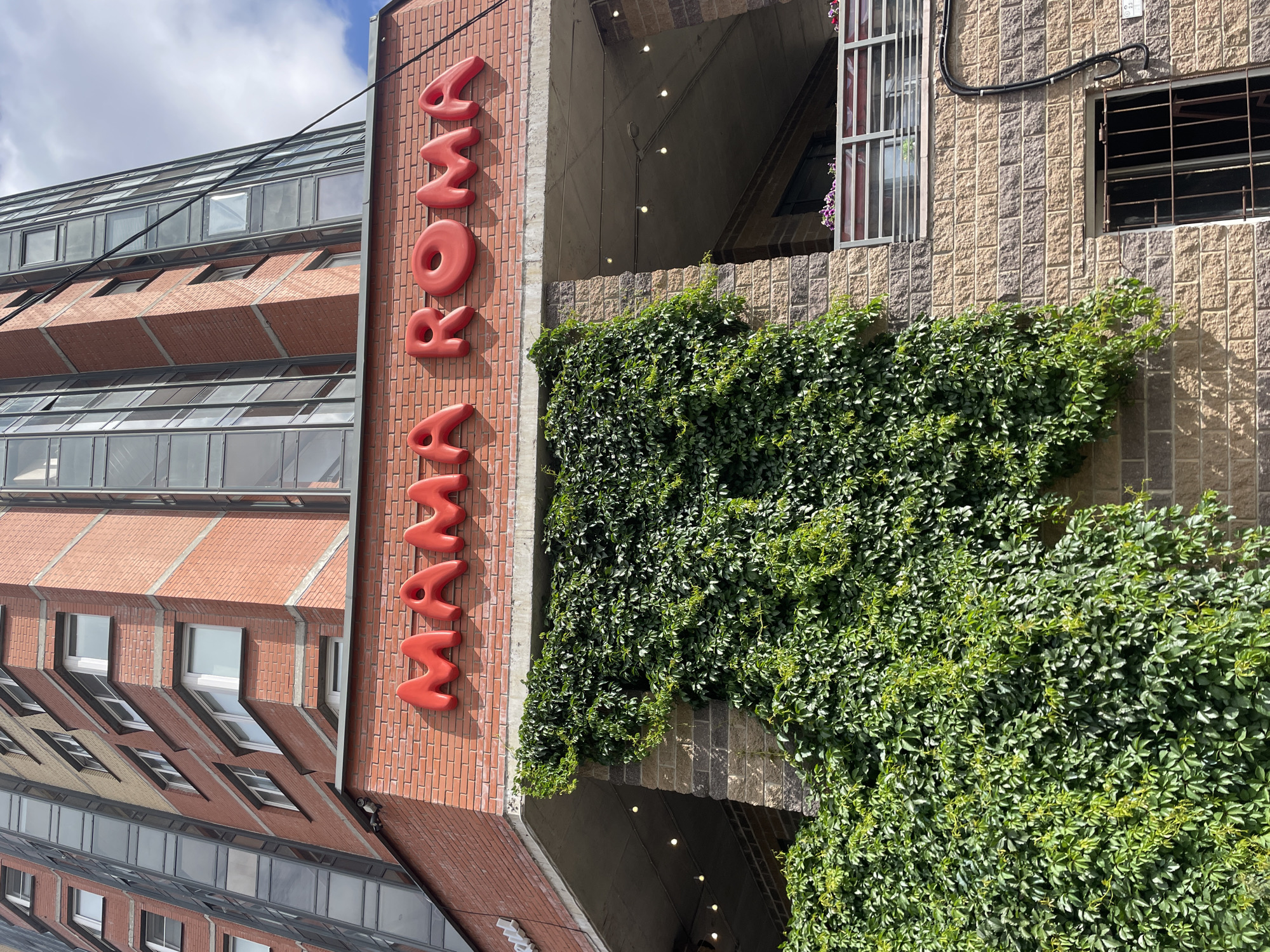 The image shows a building with red brick facade and a sign that reads "MAMA ROMA" in red, inflated letters. Green ivy covers a large portion of the lower facade, climbing up the brickwork. The building appears to be a commercial establishment. The architectural style suggests it is located in a city, possibly in Russia, given the context. The sky is partially cloudy. There are no people visible in the image.