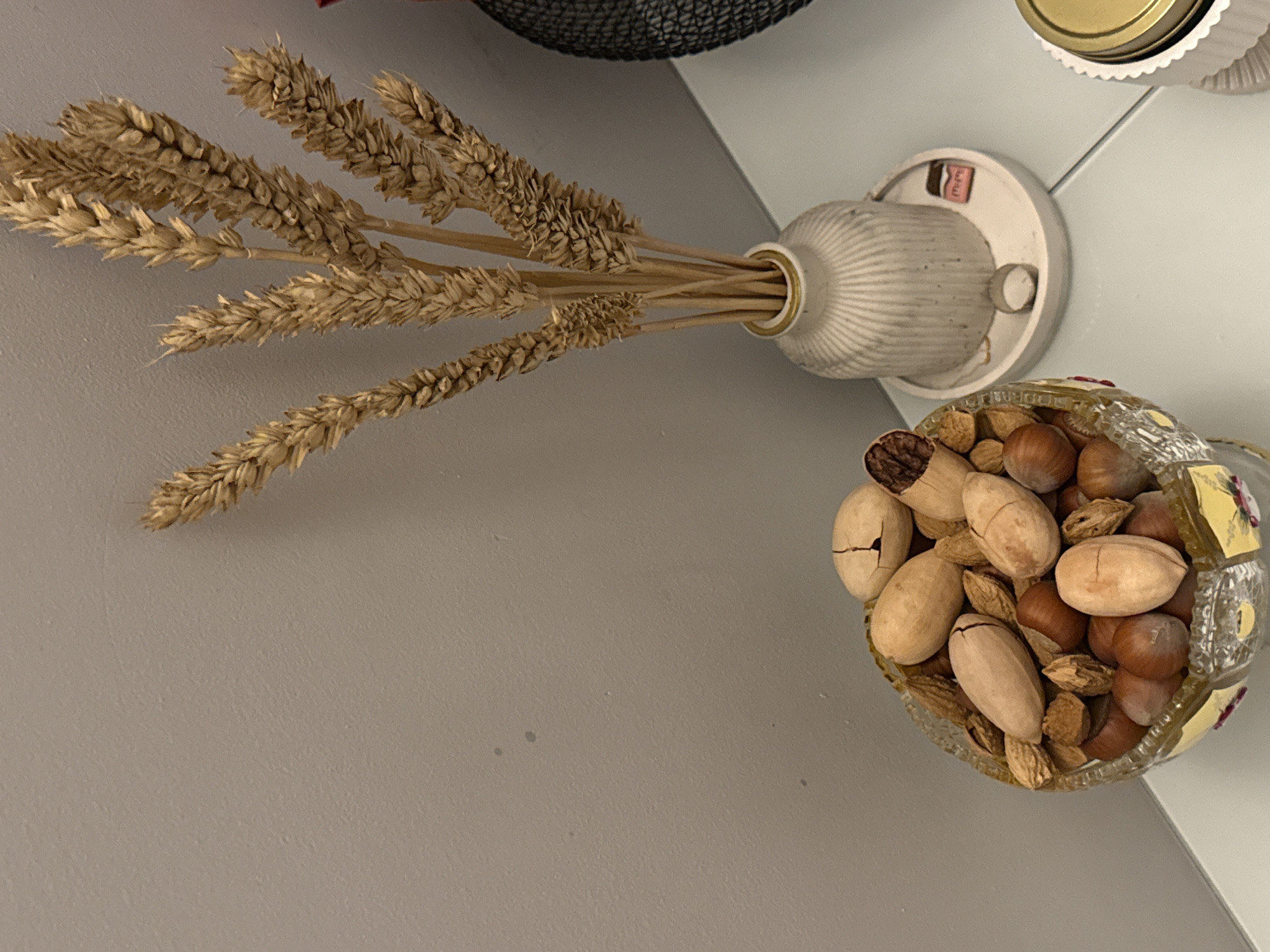 The image presents an overhead shot of a still-life composition. Arranged on a white surface that suggests a shelf or countertop, there is a collection of decorative items. In the upper-left quadrant, dried wheat stalks protrude from a textured, off-white vase mounted on a wall. To the right, a mosaic-patterned glass container overflows with assorted nuts, appearing to include pecans and hazelnuts. The backdrop transitions from a neutral wall on the left to the white surface beneath the items.