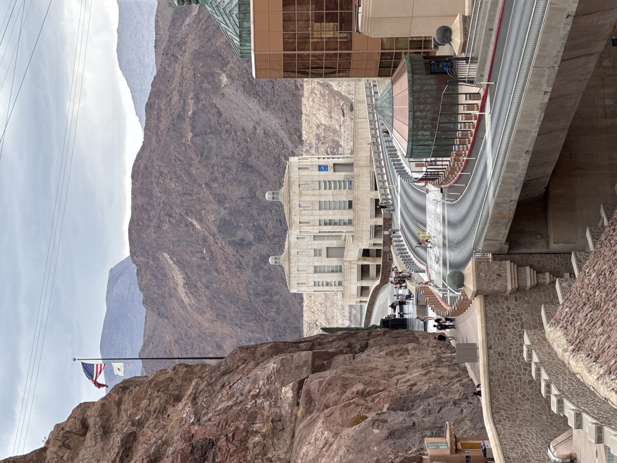 The image shows a view of the Hoover Dam near Boulder City, Nevada, on a cloudy day. In the foreground, a steep, reddish-brown rocky cliff face dominates the left side of the frame. To the right, a multi-level roadway curves through the scene, lined with modern buildings.

On the cliff face, an American flag and another white flag are prominently displayed on a flagpole. A pathway runs along the base of the cliff, with several people walking on it.

Further back, a large, symmetrical, light-colored building with a domed top and numerous windows is visible. Adjacent to it is a modern building with a copper-toned facade and a green, domed roof structure. The roadway leads toward these buildings, and there are more people visible on the pathways and the road.

The background features rugged, mountainous terrain with muted colors due to the overcast sky. Power lines are visible stretched across the sky. The overall impression is of a monumental man-made structure integrated into a dramatic natural landscape.