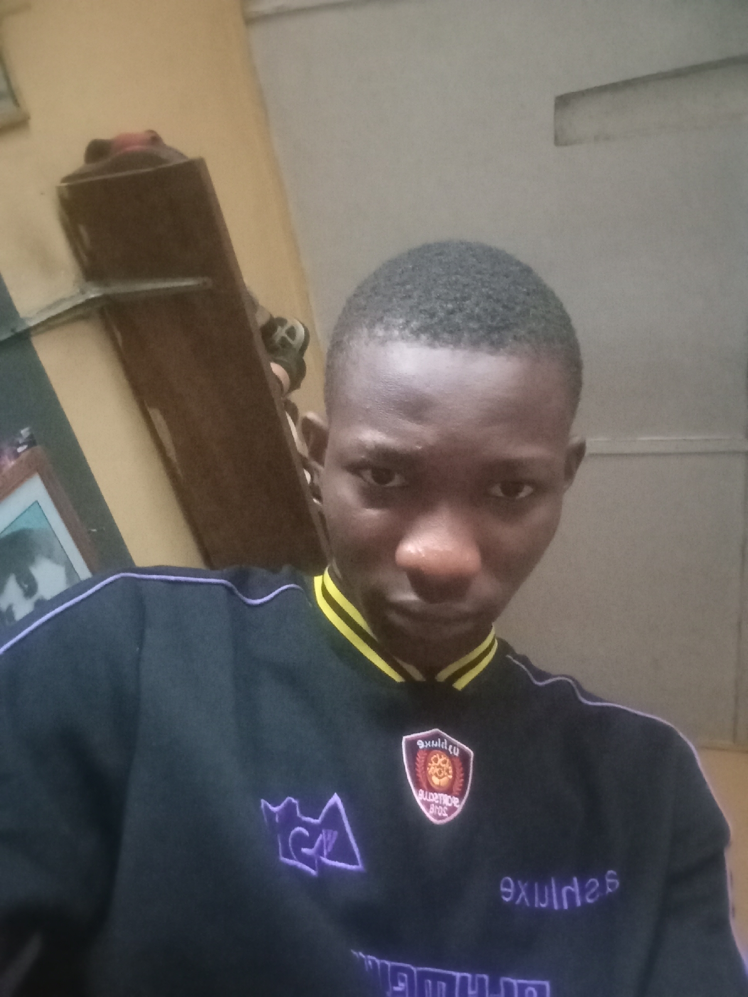 A young Black male is pictured from the chest up, looking directly at the camera. He has short, dark hair and is wearing a dark, long-sleeved shirt with yellow and purple accents. The shirt has a crest on the left side of the chest, and text on the right side. The background is a plain, light-colored wall, with a wooden shelf and a framed picture visible on the left side. The lighting appears to be from an overhead source, casting soft shadows. The scene suggests an indoor setting, possibly a home or a casual environment. The image quality is moderate, with some graininess.
