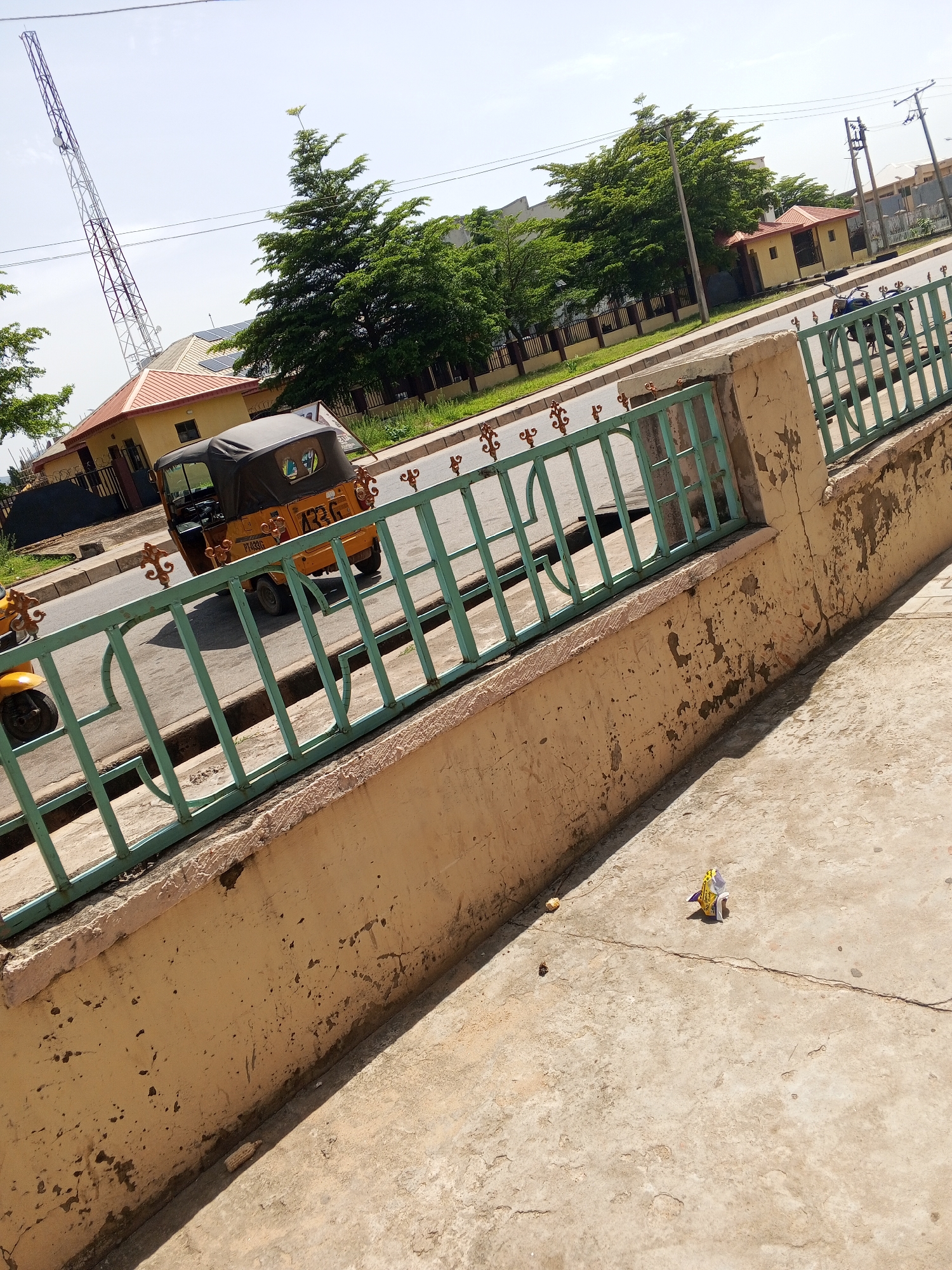 The image depicts a street scene in Jalingo, Nigeria. In the foreground, a concrete walkway is visible with a portion of a yellow concrete wall with a light green metal fence. The fence runs parallel to a road. On the road, a yellow auto-rickshaw, a common mode of transportation in Nigeria, is driving away from the camera. The auto-rickshaw has a black canvas roof and the license plate is partially visible, reading "ATZ 4220". Further down the road, several trees with lush green foliage are visible, along with buildings that appear to be residential or commercial. A tall telecommunications tower is visible in the background, a common sight in urban areas. The sky is clear and blue, indicating a sunny day. The overall impression is that of a typical urban street in Nigeria on a bright day.