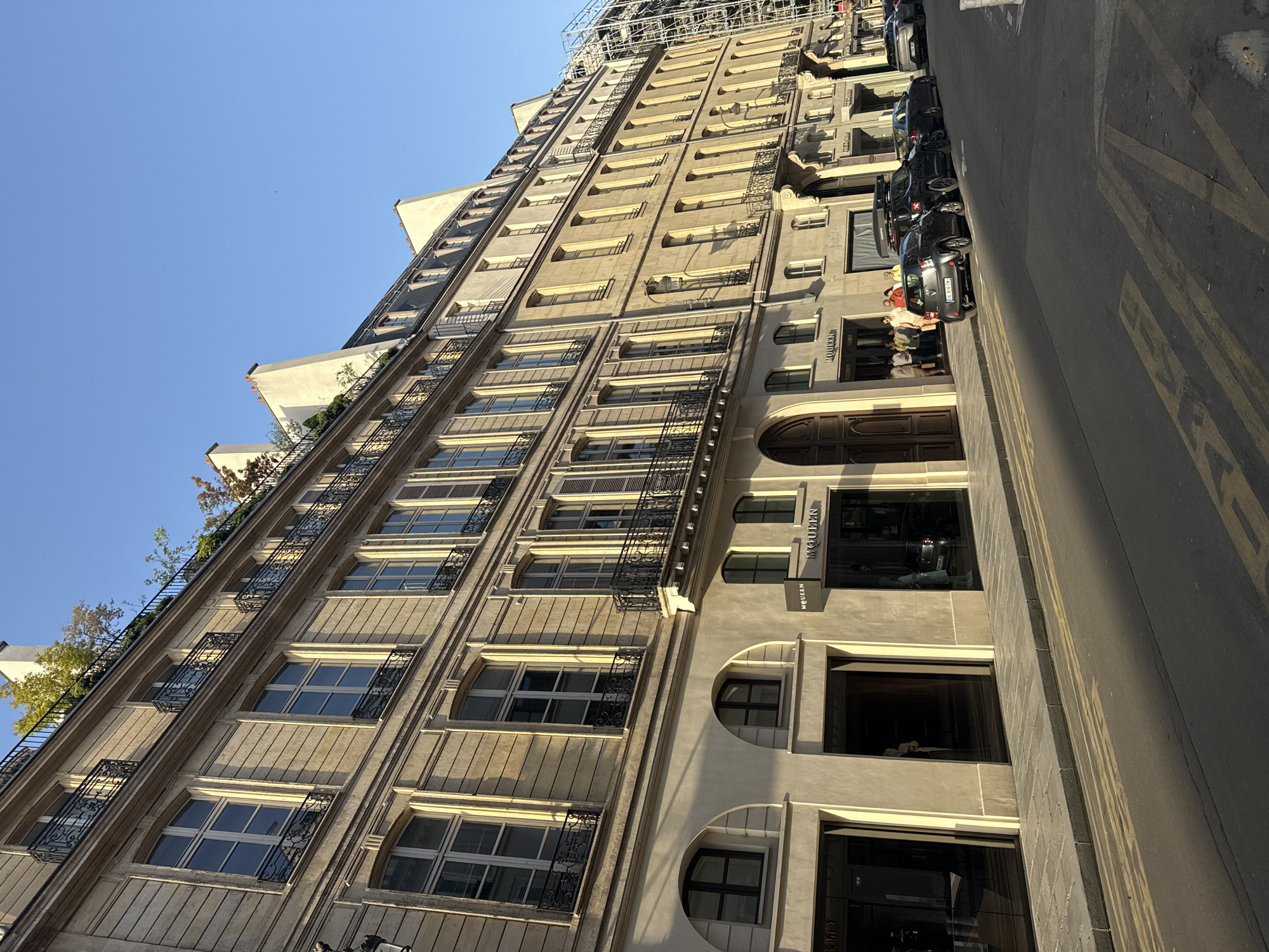The image depicts a tall, beige building in Paris, France, viewed from street level at an oblique angle. The architectural style is classic Parisian, with numerous windows, decorative iron balconies, and ornate stonework. Scaffolding is visible at the top of the building, suggesting ongoing construction or renovation.
The building houses storefronts, notably including a "McQueen" store, identifiable by signage. People are present near the store entrance. Cars are parked along the street.
The street has visible markings, including painted lines. The sky above the building is clear and blue. The scene is bathed in sunlight, casting shadows across the building's facade and the street.