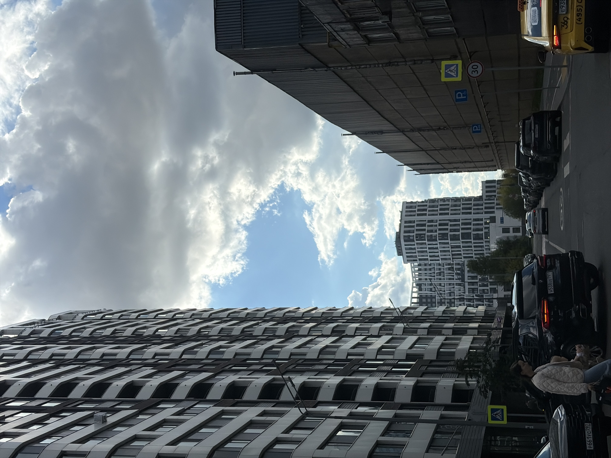An exterior, eye-level, medium shot captures a street scene in Moscow, Russia, under a cloudy sky. On the left, a modern, tall residential building with a dark and light patterned facade dominates the view. To the right, a long, industrial-style building with a corrugated metal exterior recedes into the background, with a lower, older building in front of it. In the middle ground, several cars are parked and driving on the road. A black SUV with illuminated taillights is prominently featured, and a yellow taxi is visible on the far right. A woman and child are walking on the sidewalk in the foreground, with a pedestrian crossing sign visible. The overall impression is of an urban environment with a mix of contemporary and older architecture, indicative of ongoing development and existing infrastructure within the city.