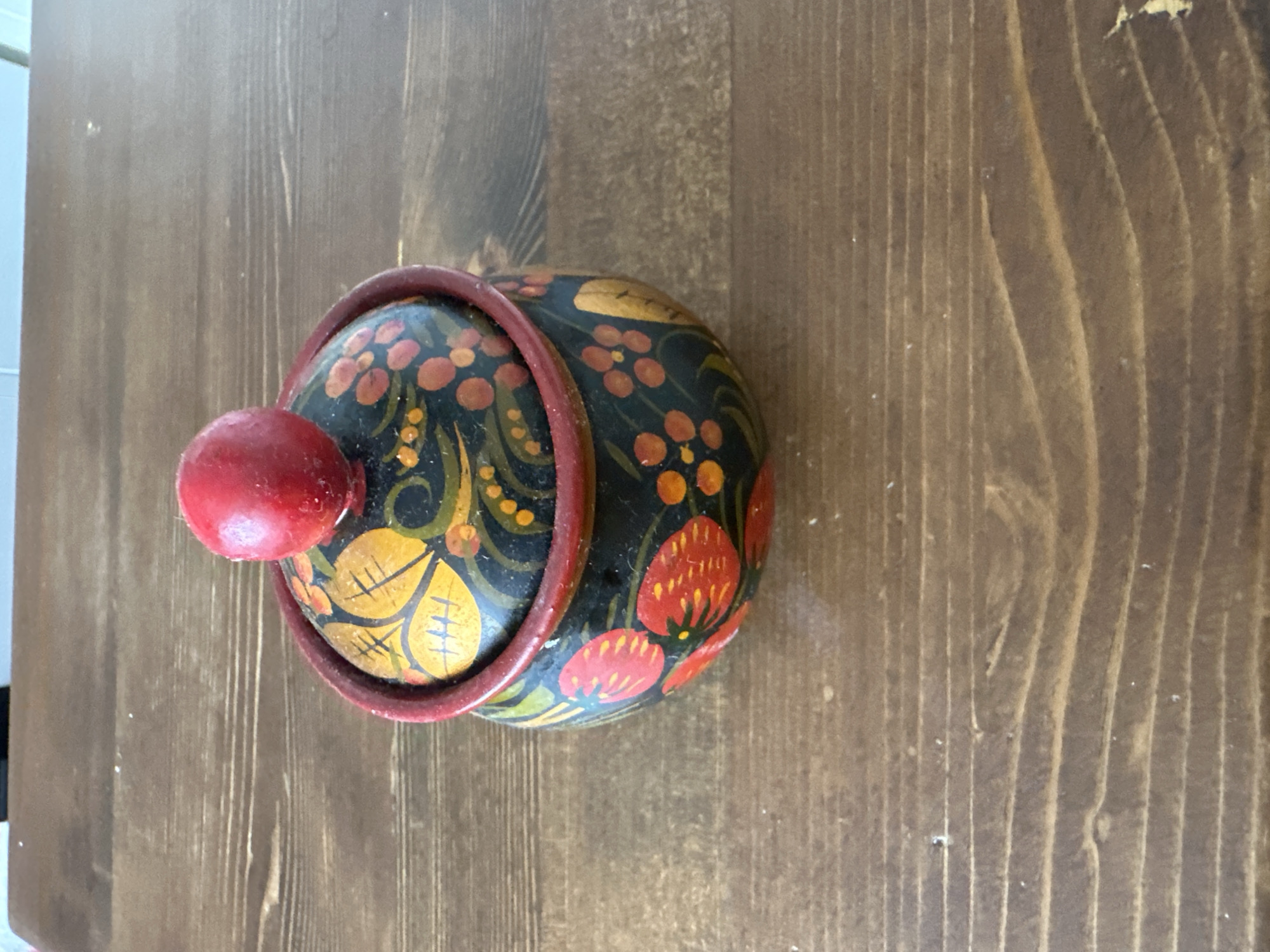 An overhead shot shows a wooden pot with a lid on a wooden table. The pot is decorated with a black background and colorful floral patterns in red, yellow, and orange. A red, spherical knob sits on top of the lid. The table is made of dark wood planks with visible grain. The background is blurred, showing hints of a lighter colored wall or surface. The scene is static and appears to be indoors. The image is in focus and well-lit. This scene is not in Balqaş, Kazakhstan.