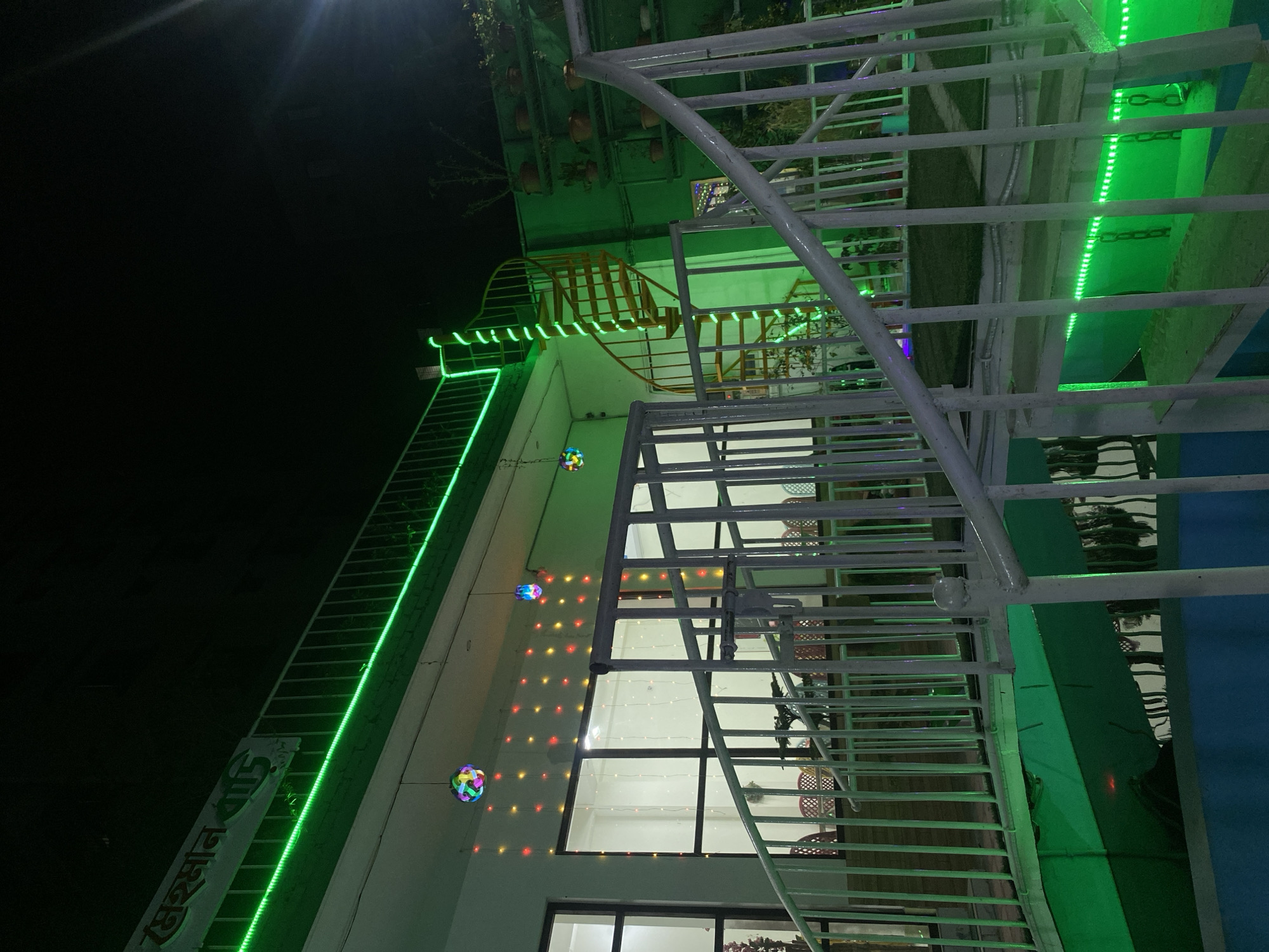 Captured at night in Dhaka, Bangladesh, the scene depicts the exterior of a building adorned with vibrant, colorful lights. A metal staircase dominates the foreground, its bars casting linear shadows. In the background, green LED lights outline the building's edges, and a spiral staircase is visible. Strings of decorative lights hang along the wall of the structure. The lighting and composition suggest a festive or celebratory atmosphere. A sign with Bengali script is also visible, illuminated with green lights.