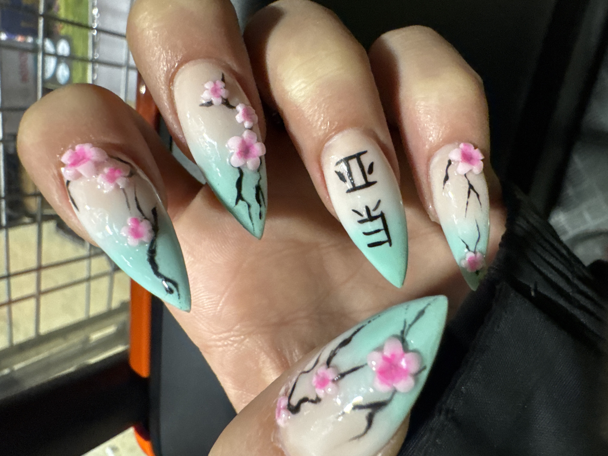 A close-up image shows a person's hand with manicured nails, featuring a white and pale blue gradient design. The nails are pointed. The designs include hand-painted black branches and pink cherry blossom flowers. One nail has two vertical black characters painted on it. The background is out of focus, but includes a metallic structure and a dark garment. The location, Kettering, United Kingdom is not discernible within the image.