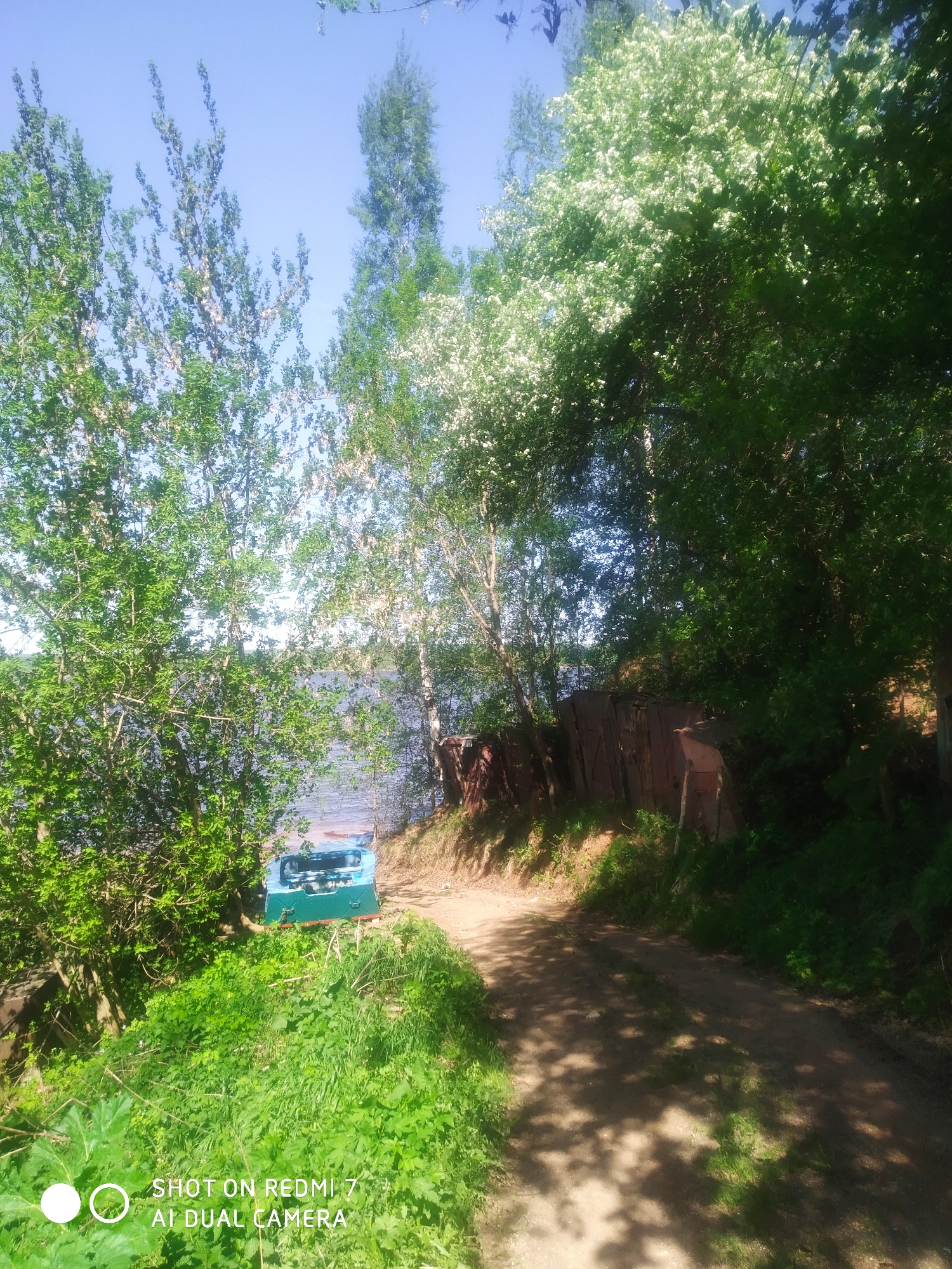 The photograph, taken in Ochansk, Russia, presents a landscape scene featuring a dirt path winding along a shoreline. The path is dappled with sunlight filtering through dense foliage. To the left of the path, a cluster of trees leads down to a body of water, with a small, overturned blue and green boat situated among the greenery. Adjacent to the path on the right is a densely wooded area with what appears to be a rusted corrugated metal structure partially obscured by the trees and foliage. The sky is visible through the trees. The lower portion of the image includes a patch of vibrant green vegetation.