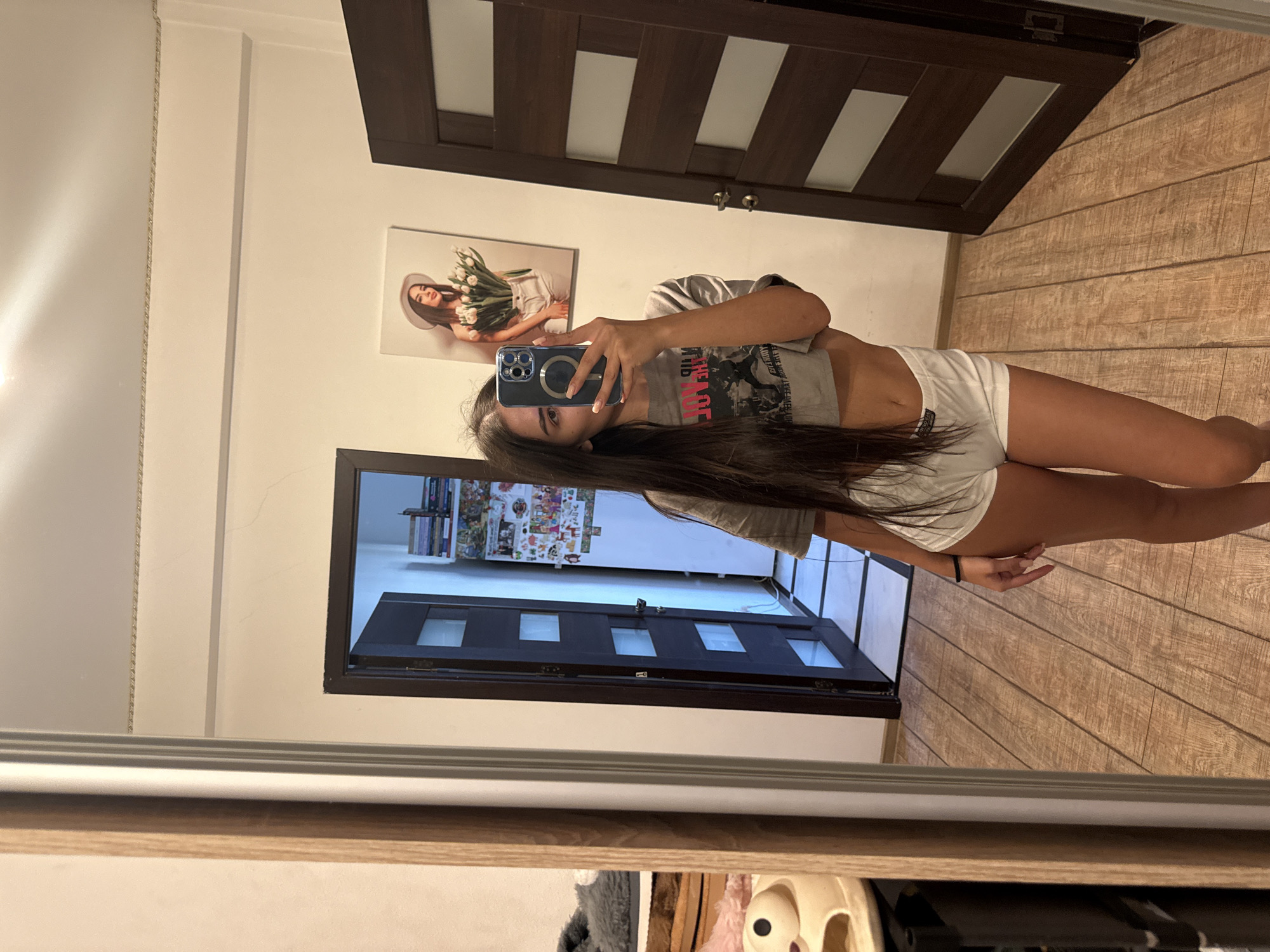 The image shows a young woman taking a mirror selfie in what appears to be a home interior. She is wearing a grey crop top with text graphics and white shorts. The setting includes a wooden floor, a dark wooden door with glass panes, and a picture on the wall of a woman with tulips. A bookshelf is visible in the reflection of the mirror, showing various books and decorative items. The woman is holding a smartphone, likely to capture the selfie. The overall mood is casual and relaxed, typical of a personal photograph. There is no indication of the time of day or weather.