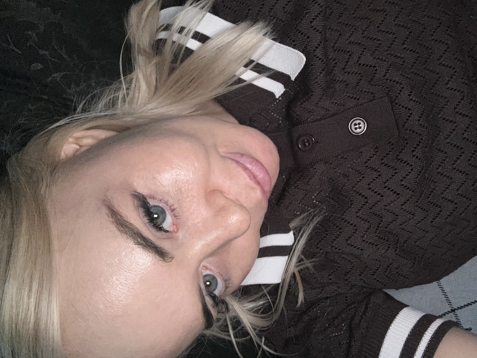 A fair-skinned female individual with blonde hair and blue/grey eyes is depicted in a close-up, self-portrait style image. She is looking directly at the camera with a subtle expression and slightly parted lips. Her hair is light blonde, with some strands loose around her face and ear. Her eyebrows are dark and defined, and she wears dark winged eyeliner and long dark eyelashes. A small reddish irritation is visible on the lower eyelid margin of her left eye (viewer's right). Her skin shows visible texture, particularly on the forehead, and appears somewhat reflective in areas such as the forehead, nose, and cheekbone.
She is wearing a dark brown, knitted polo-style shirt with a white collar featuring horizontal brown stripes. The shirt fabric has a distinctive openwork chevron or zigzag pattern. Two dark circular buttons are visible on the placket. The short sleeves also feature white and brown striped cuffs.
The background directly behind her head and right shoulder is dark and indistinct, possibly a dark wall or fabric. To the lower left, beneath her left arm and sleeve, a light blue/grey fabric with a faint diamond or argyle pattern is visible. The lighting appears artificial, casting highlights on her skin, and the overall environment is dimly lit. The location is in the city of Unknown, Unknown.