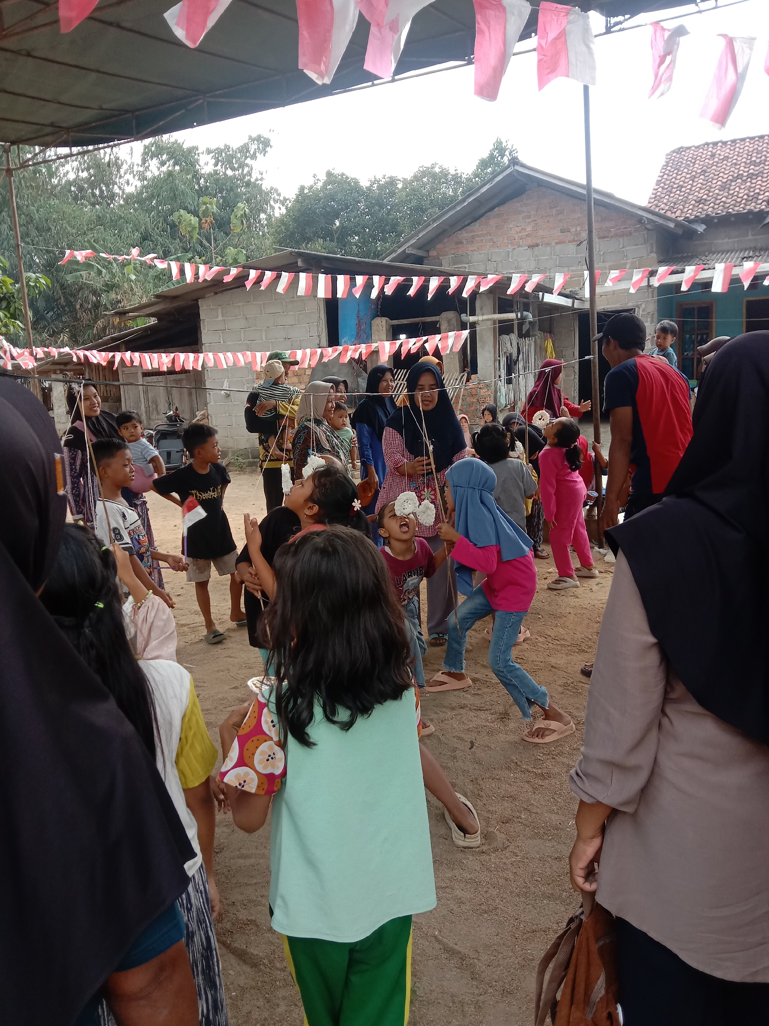 The image captures an outdoor event, likely a celebration or competition, in Utama Jaya Mataram, Indonesia, as evidenced by the Indonesian flag colors decorating the scene. A group of people, primarily children, are engaged in a game. Several children are attempting to bite onto pieces of food suspended from strings. The environment appears to be a dirt-grounded area with rudimentary structures and trees in the background, suggesting a village or less developed urban setting. Red and white Indonesian flags are strung across the area, indicating a national celebration. The people are dressed casually, with some wearing headscarves. The overall atmosphere seems lively and festive.
