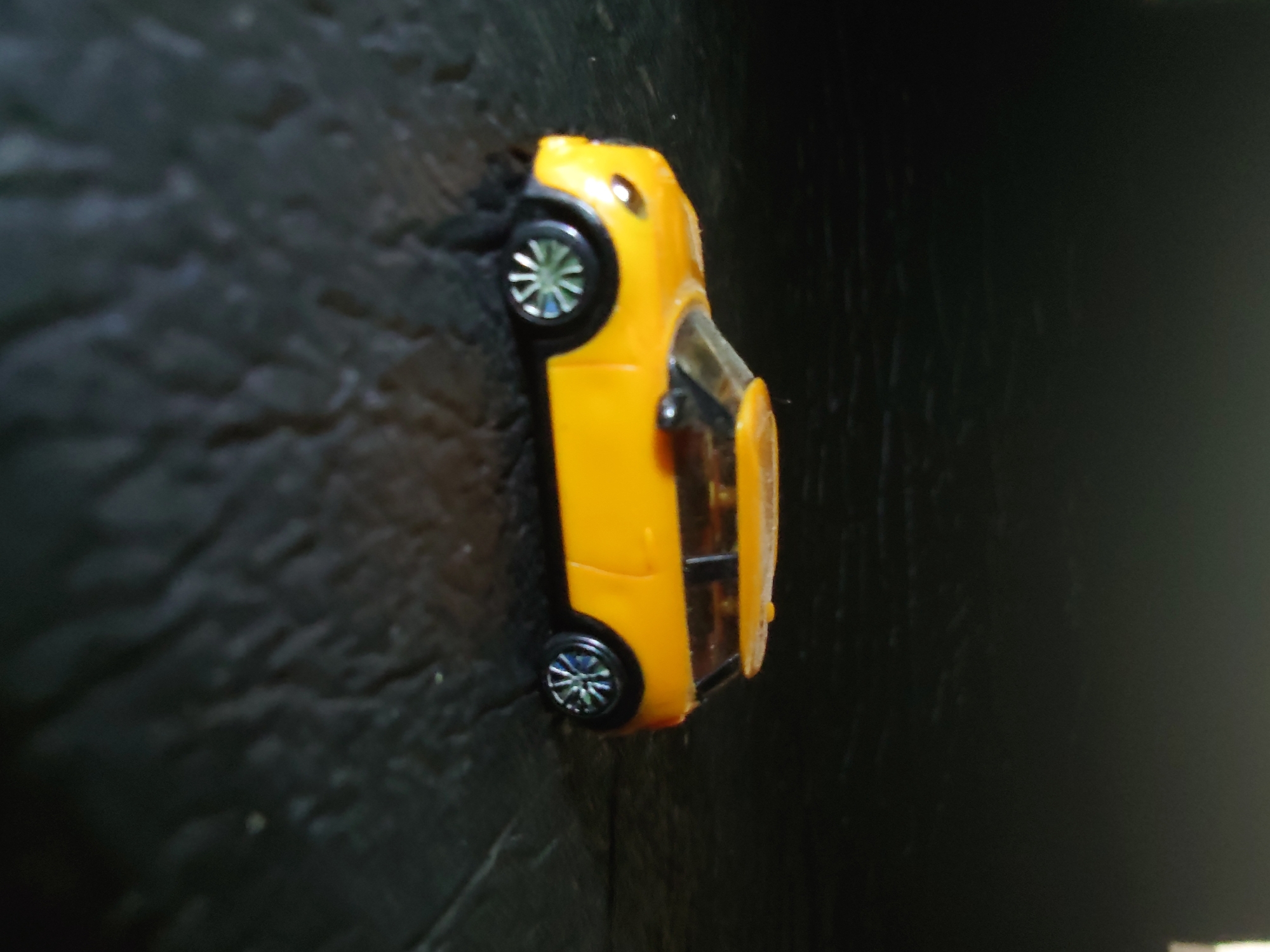 A yellow toy car, resembling a Mini Cooper, is positioned on a dark, textured surface. The car is facing left, with its front headlights visible. The wheels are black with silver rims. The background is dark and out of focus, with a slight sheen suggesting a reflective surface. There is no indication of the car being in motion or any interaction with other objects or individuals. The scene appears to be an indoor or outdoor setting with minimal ambient light. There are no people present in the image. The overall composition is simple, focusing solely on the toy car as the main subject. The image does not provide any geographical context related to Sokolohirsk, Ukraine.