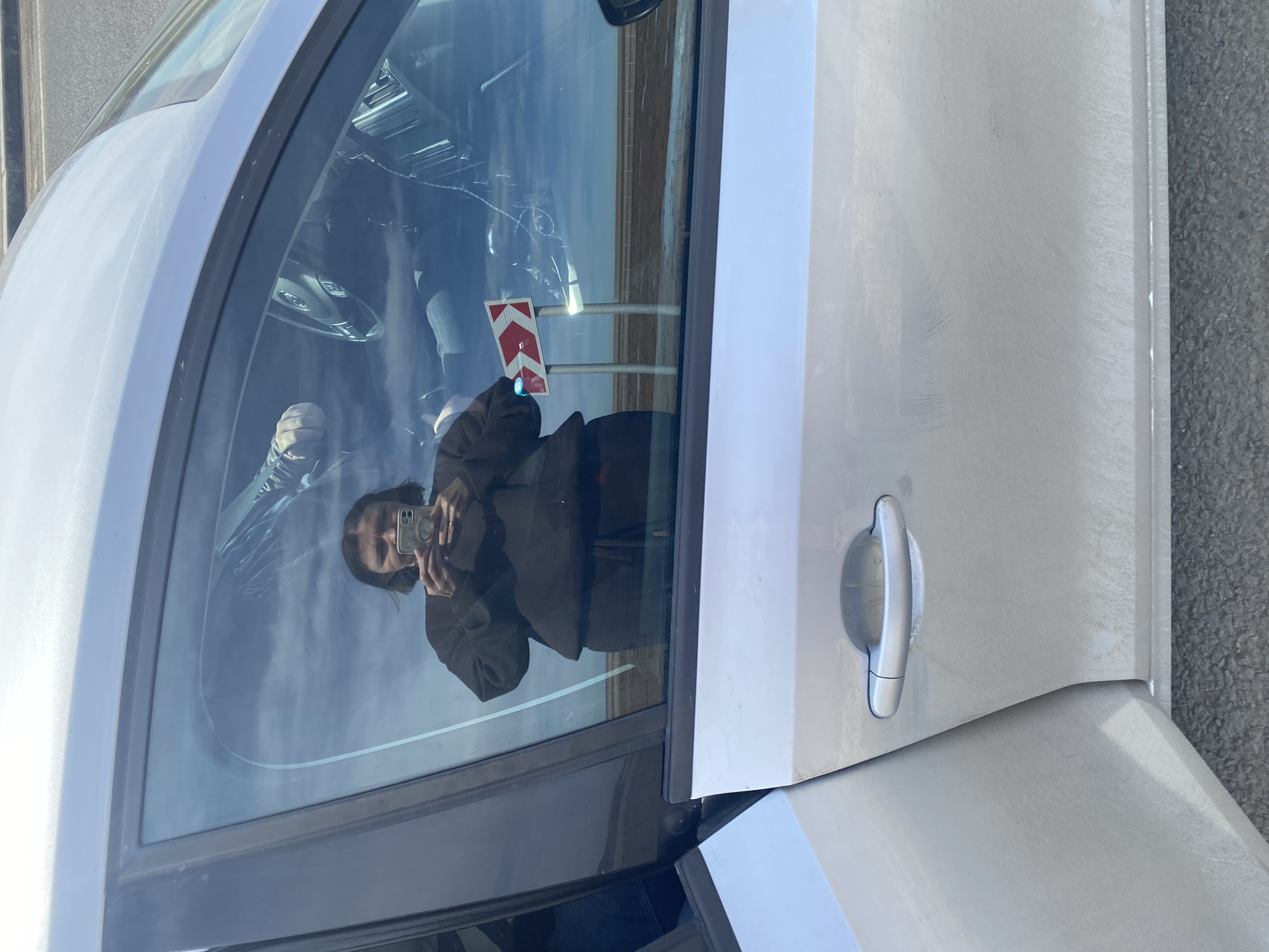 Here's a detailed description of the image:
**Scene:** The image shows the door of a silver-colored car. A person, presumably the photographer, is reflected in the car window, holding a smartphone and taking a picture. The background visible through the window suggests an outdoor environment.
**People:** One person is visible as a reflection. Their appearance is partially obscured by the reflection, but they appear to be holding a smartphone.
**Objects:** Key objects include the silver car, specifically its door, the car window, a smartphone held by the reflected person, a road sign.
**Environment:** The road sign, reflecting the light, appears to be in Kliuchiki, Russia. The sign indicates a direction and is along a roadside.