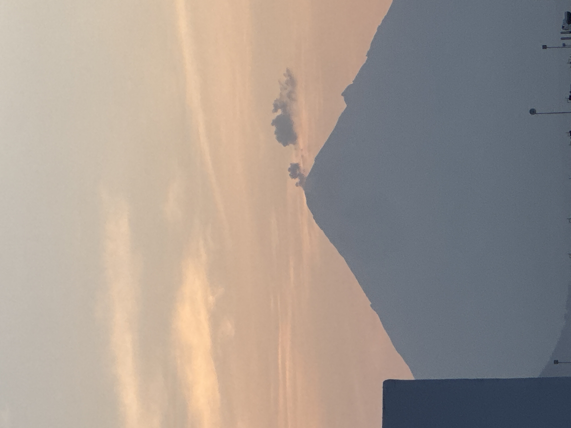 The image depicts a mountain range near San Francisco Ocotlán, Mexico, under a sky with wispy, light-colored clouds tinged with hues of orange and pink, suggesting either sunrise or sunset. A darker, cumuliform cloud is located near the peak of one of the mountains. Along the mountain range are a series of utility poles with lights. In the lower portion of the image is a dark rectangular shape that appears to be the top of a building or structure.