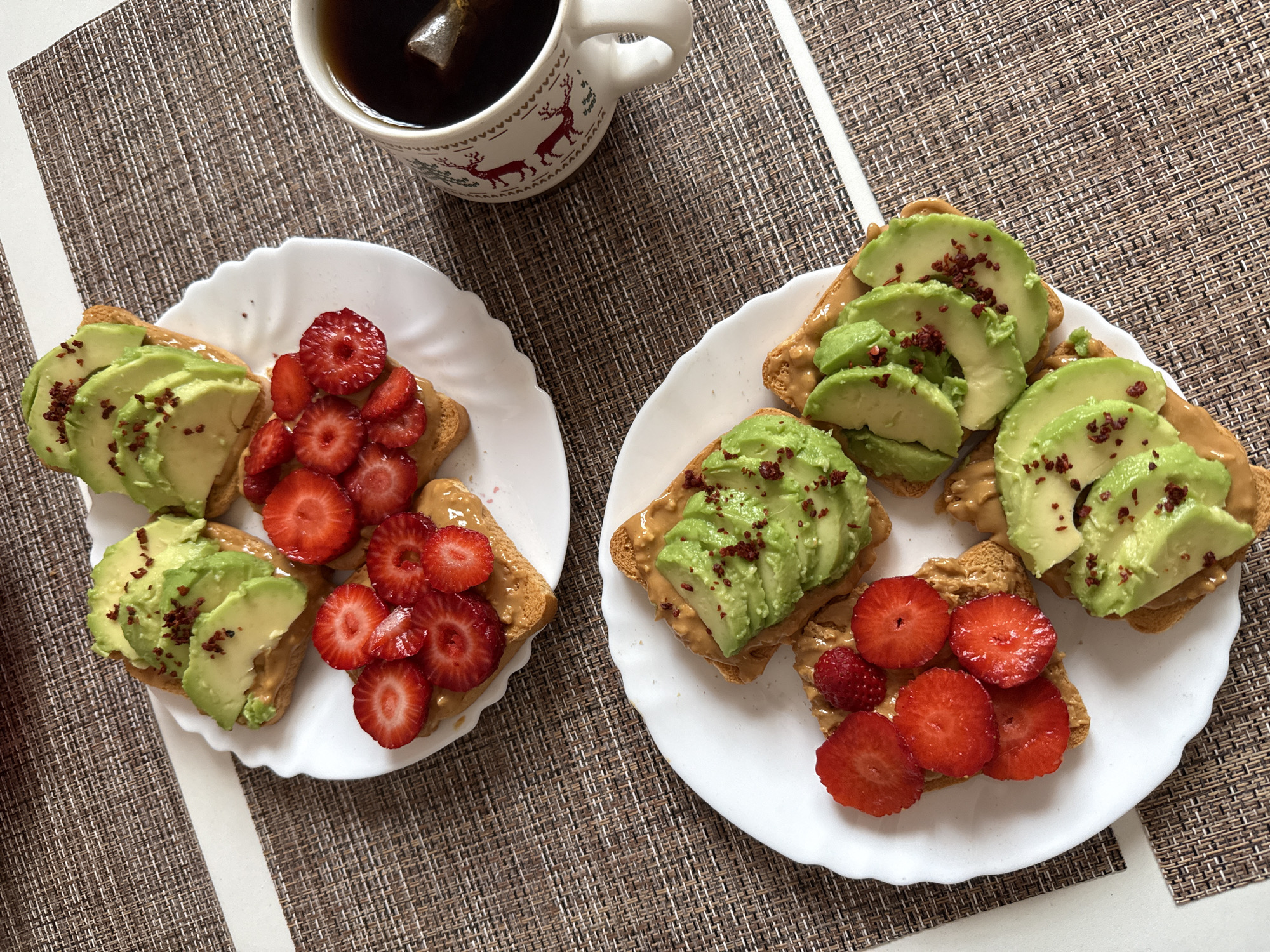 Two white plates, each holding four slices of toast topped with peanut butter, are arranged on a woven placemat. One plate features toast topped with sliced strawberries and a single raspberry, while the other has toast topped with sliced avocado sprinkled with red pepper flakes. A white mug with a deer pattern, containing dark liquid and a tea bag, sits to the right of the plates on the placemat. The background is a flat surface, possibly a table, with a white edge visible. The scene appears to be indoors, likely in a domestic setting, and there is no indication of a specific city or location.