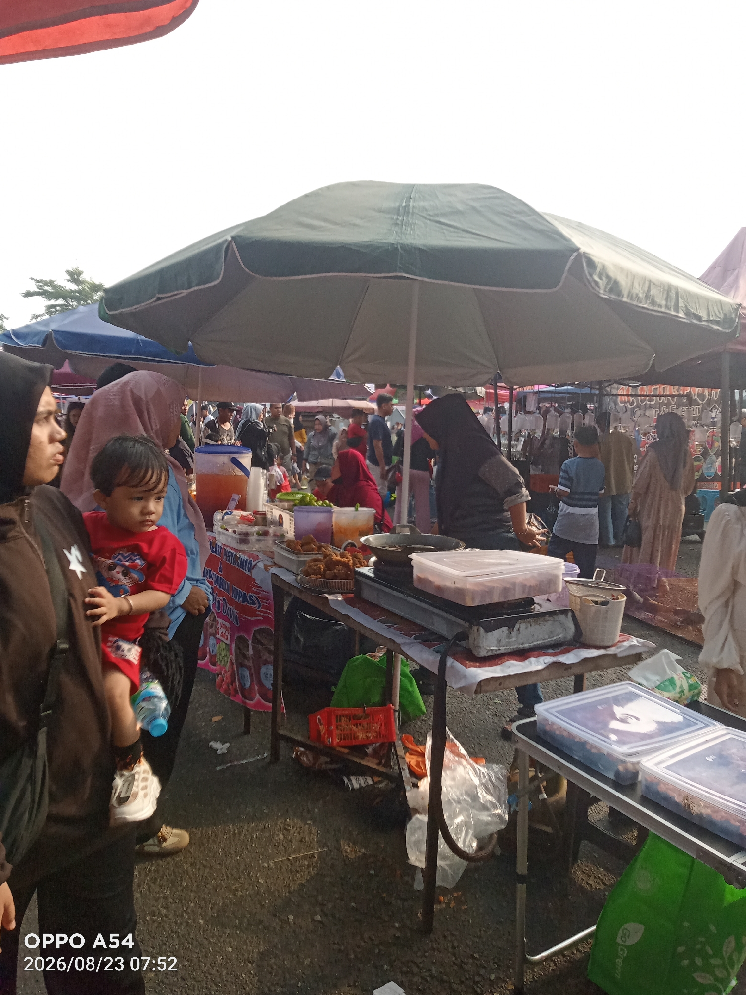 The image shows an outdoor market scene, likely in Jambi, Indonesia, as indicated by the timestamp. Several large umbrellas provide shade over stalls. In the foreground, a woman wearing a dark headscarf holds a young child in a red shirt. To their right, another woman in a light pink headscarf is visible. The stalls display various food items, including fried goods, drinks in a large dispenser, and food in plastic containers. People are browsing and interacting, suggesting a lively market atmosphere. The ground appears to be asphalt, common in urban areas. The lighting suggests it is daytime, possibly morning given the timestamp.