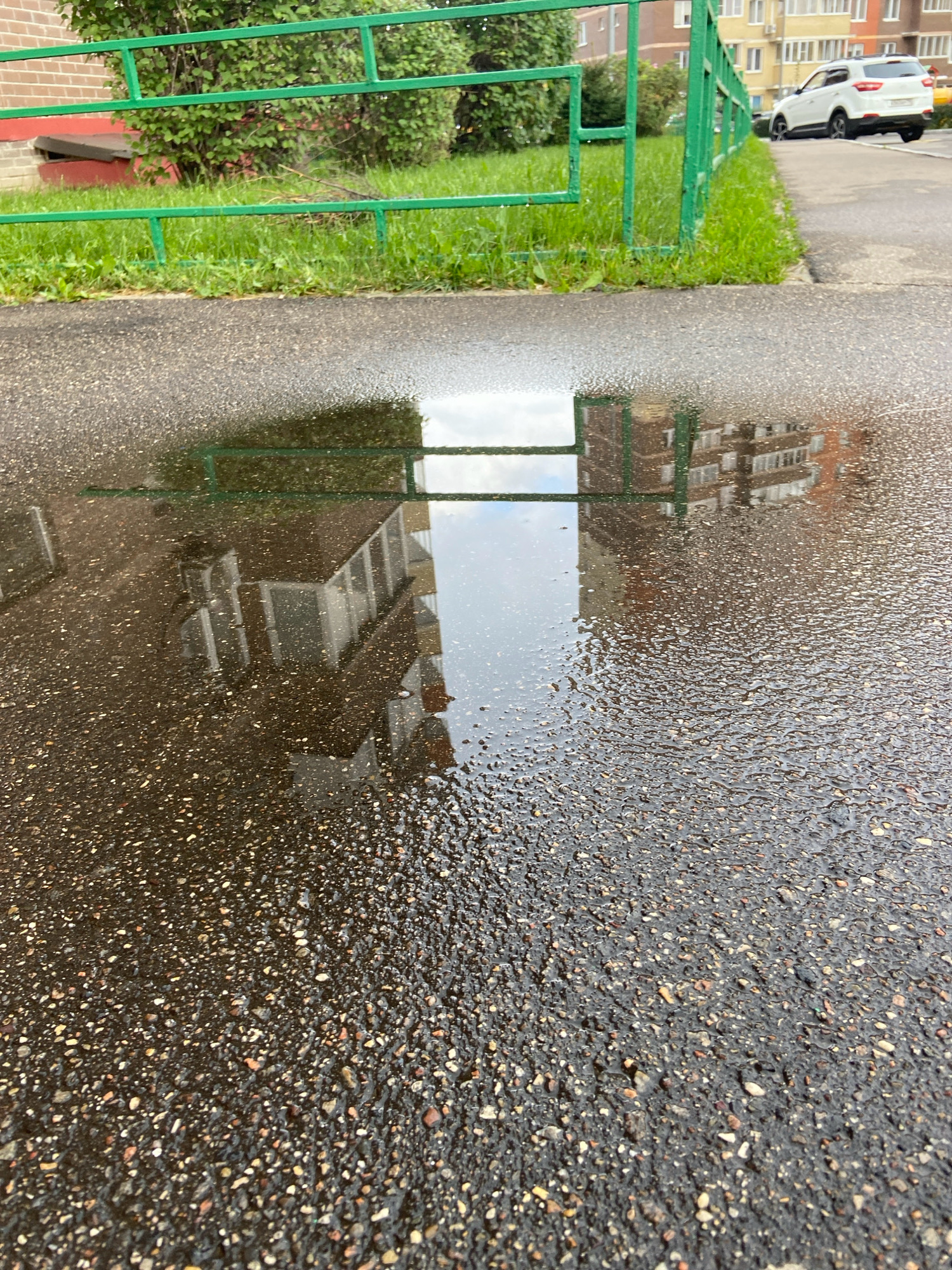 An overhead, eye-level, wide-angle shot captures a puddle on asphalt in Balashikha, Russia. A green metal fence runs horizontally across the top of the image, bordered by grass and a red brick building on the left, and a sidewalk with a white SUV on the right. The puddle reflects a multi-story brick building and the sky, its surface textured with ripples and droplets. The asphalt itself is dark and speckled with small stones.