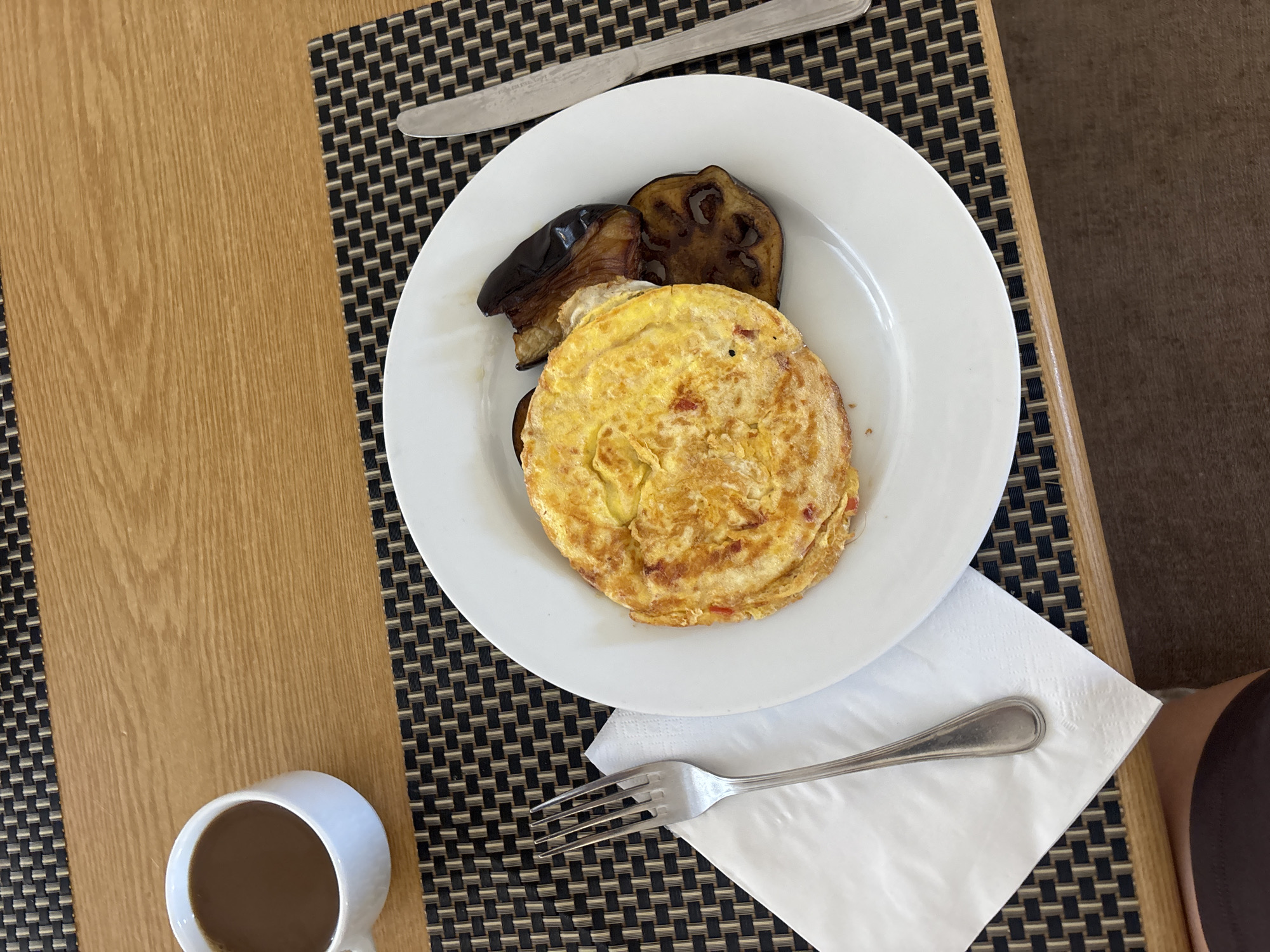 A top-down shot depicts a meal setting on a table, featuring a white plate with an omelet and eggplant slices. The plate rests on a white napkin, positioned on a dark brown woven placemat. A fork is placed on the napkin, and a knife is set above the plate on the placemat. A white mug with a dark liquid, likely coffee, sits to the left of the plate. The table appears to be made of wood, with one section light brown and the other a darker shade. The location is Unknown, Unknown.