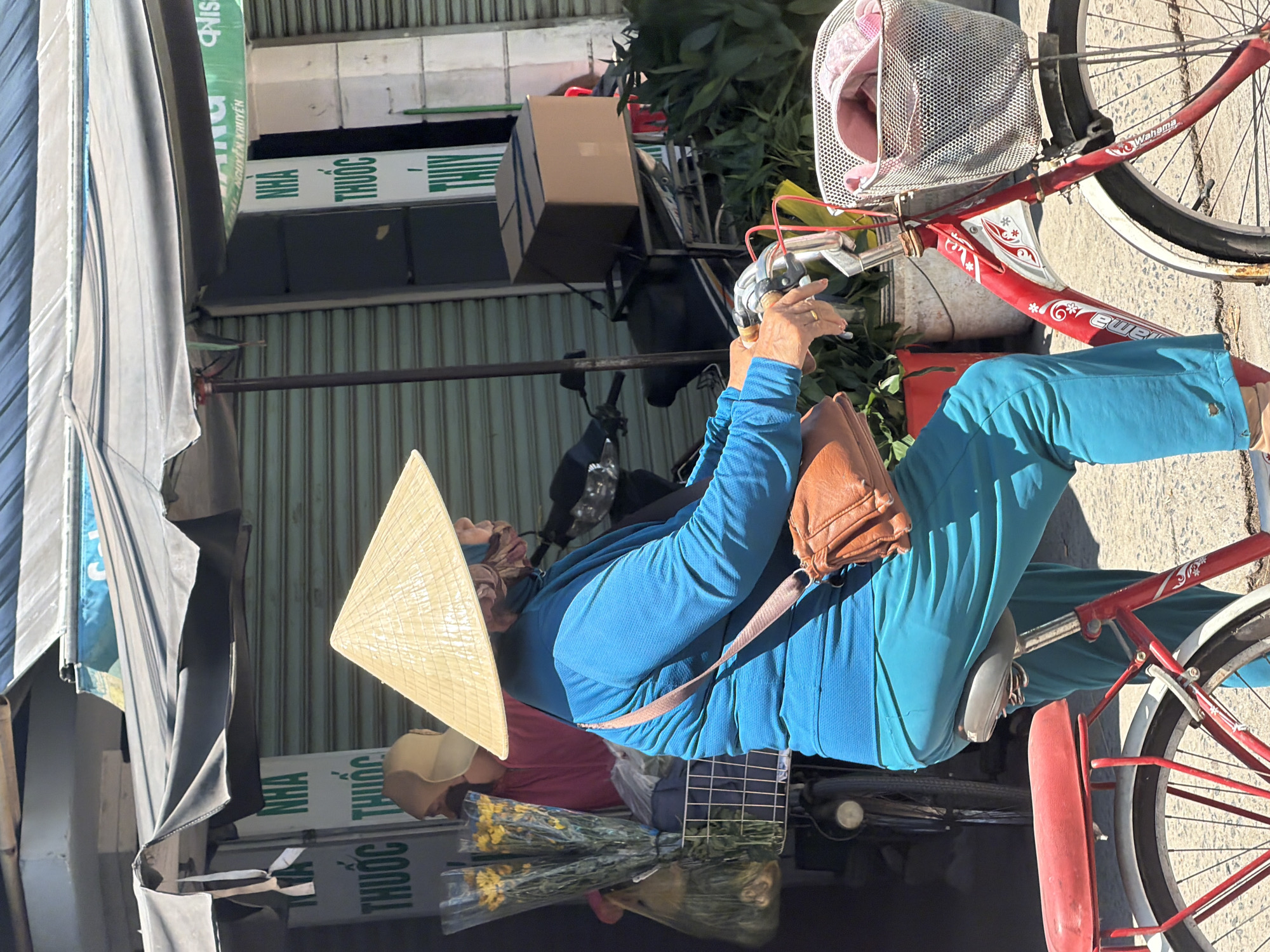 The image shows a person wearing a conical hat and a blue shirt riding a red bicycle on a street in Bac Nha Trang Ward, Vietnam. The person is carrying a brown shoulder bag. The bicycle has a basket in the front containing a pink bag. In the background, there is a store with green shutters and signs in Vietnamese. There are also plants and a cardboard box visible. The lighting suggests it is daytime.