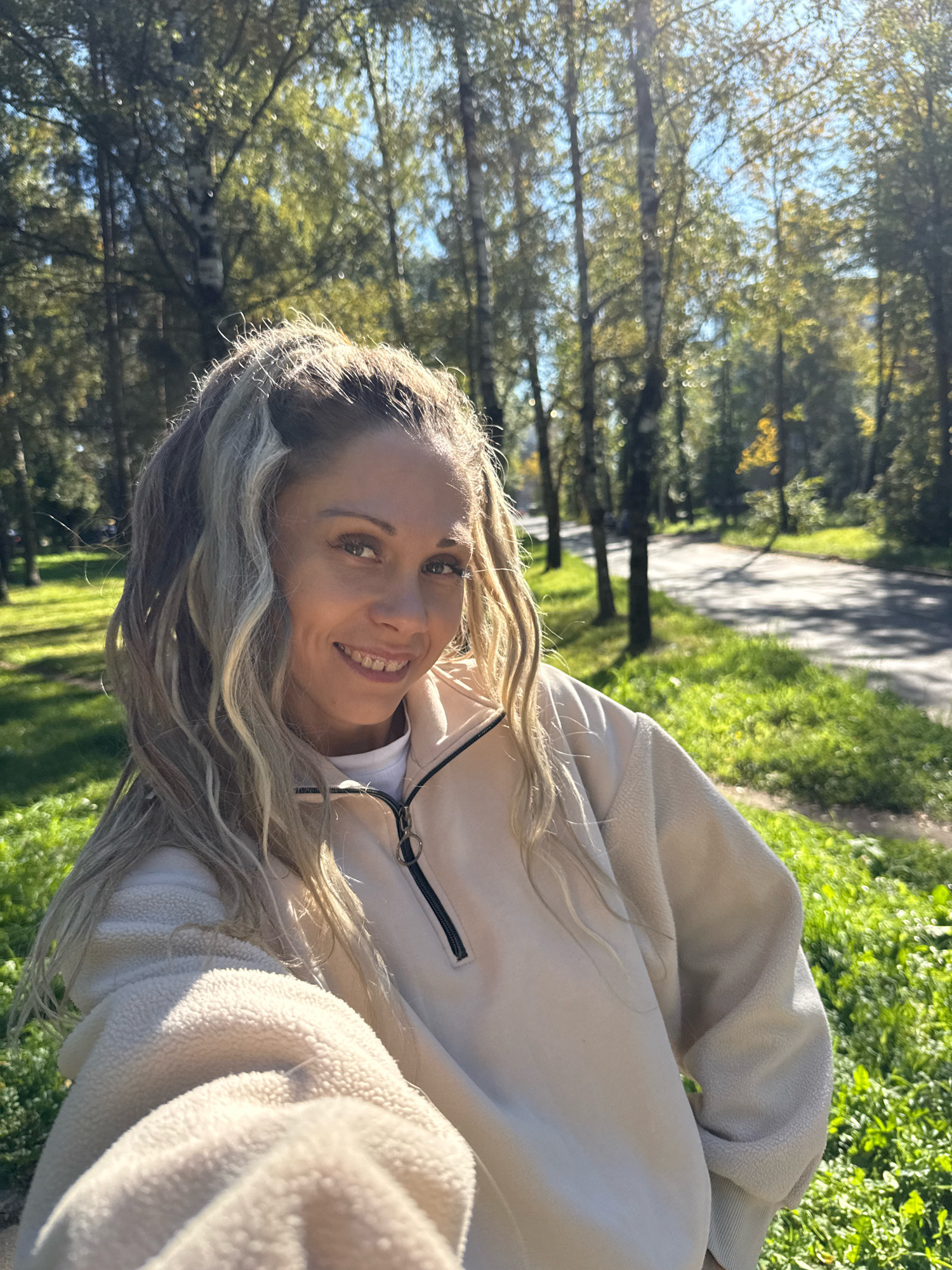 A woman with blonde hair is taking a selfie outdoors in a park. She is wearing a cream-colored fleece jacket with a zipper. The background shows a path or road lined with trees and green grass. The sun is shining, casting shadows and creating a bright, sunny atmosphere. The location is specified as Rusanovo, Russia.