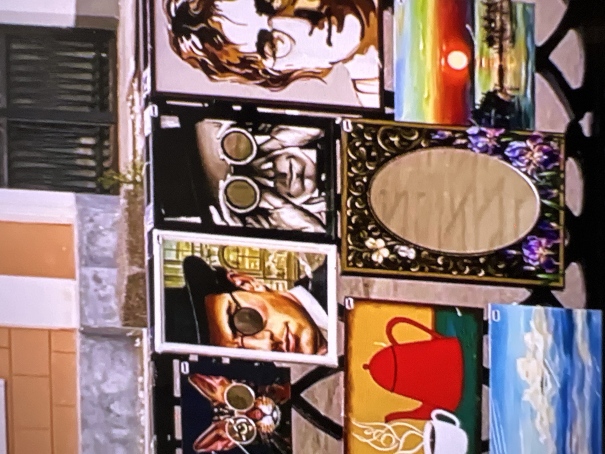 This image displays a collection of framed artworks, possibly arranged for sale or exhibition. The subjects vary widely, including portraits of people, still life compositions, and abstract or landscape elements.
In the upper portion of the image, a portrait of a person with distinctive swirling brown and white hair is visible. Below this, another portrait features a person wearing goggles, with two circular lenses prominently displayed. To the right of these portraits, a piece depicts a sunset with a rainbow-like gradient of colors and a silhouetted forest scene.
Further down, a framed mirror with an ornate, dark frame and floral accents is positioned centrally. Below the mirror, another portrait shows a person wearing a hat and goggles, with a golden-toned background. To the right of this portrait, a still life painting features a bright red teapot and a white mug on a yellow background. Adjacent to this is a painting with blue and white strokes, suggesting clouds or water. At the bottom, a framed image appears to be a stylized depiction of a cat's face, also with goggles.
There is no indication of specific activities or events taking place. The setting appears to be indoors or in a sheltered outdoor space, given the arrangement of framed items. Location cues like windows with horizontal blinds are visible on the left. No specific time of day, weather conditions, or emotional cues are discernible from the artworks themselves. The number "10" is visible on one of the artworks.