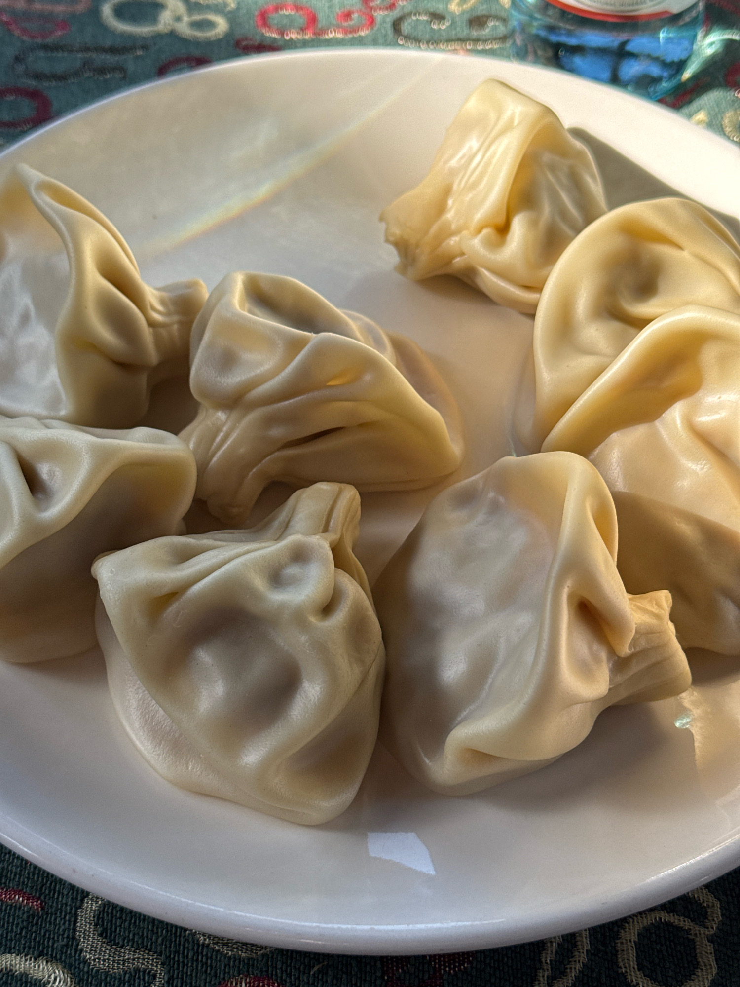 A close-up, overhead shot shows a white plate of khinkali, traditional Georgian dumplings. The dumplings are light yellow, plump, and gathered at the top with a twisted knot. They are arranged on the plate in a somewhat haphazard fashion, with some overlapping. The plate is slightly angled, allowing the viewer to see the curved edge of the plate and a hint of the patterned tablecloth underneath. A faint rainbow-like reflection is visible on the plate, possibly from sunlight. In the upper right corner, a portion of a blue bottle with a white cap is visible, suggesting a beverage is also present. The lighting casts shadows, highlighting the texture and form of the khinkali. The overall scene is likely from a restaurant or home in Telavi, Georgia, a region renowned for its cuisine.