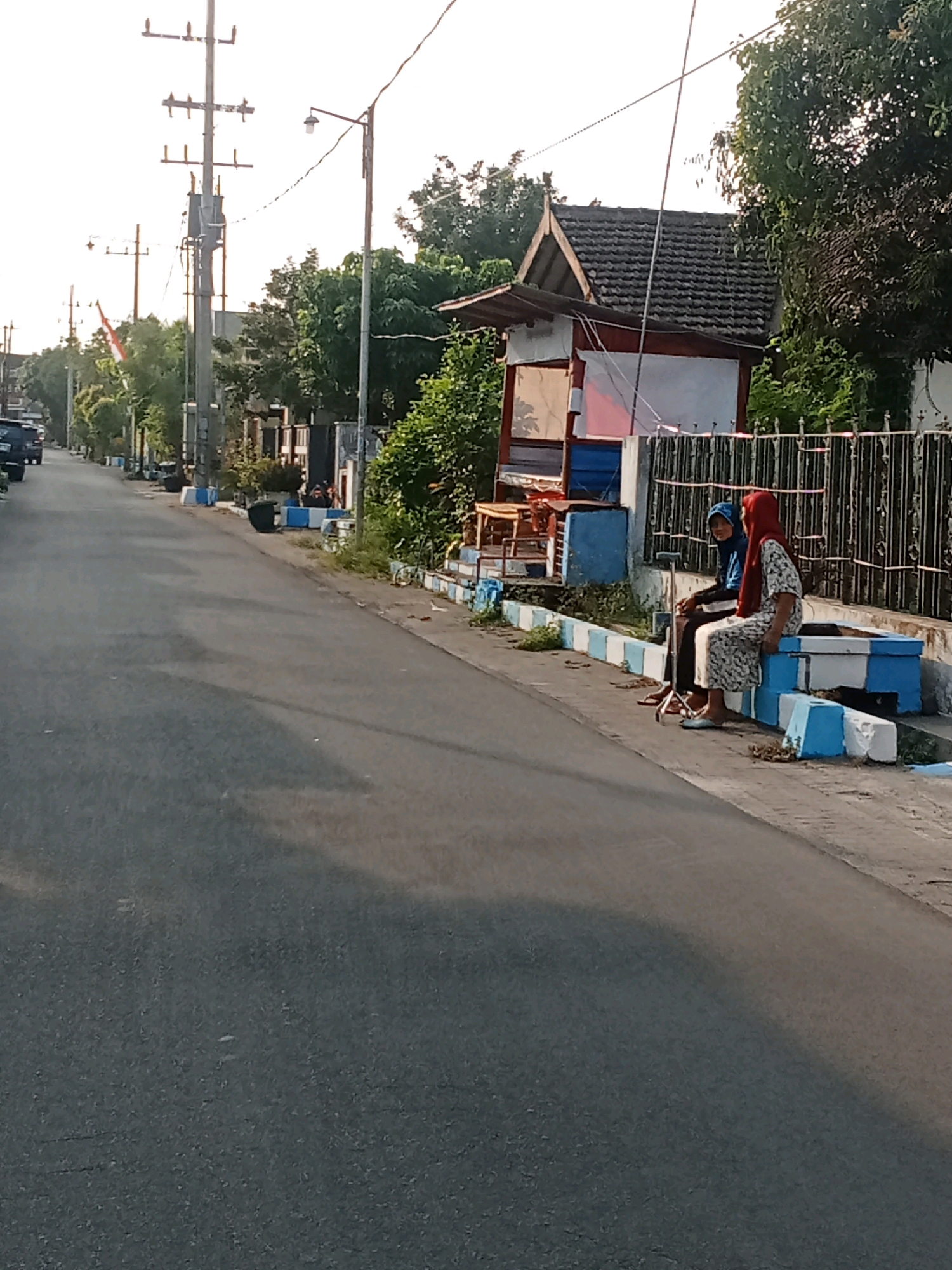 In the city of Kaliwungu, Indonesia, a paved road stretches into the distance under a bright sky. On the right side of the road, two individuals are seated on a concrete structure painted with white and blue blocks. The person on the left wears a blue headscarf and dark clothing, while the person on the right has on a red headscarf and a patterned dress. Next to them, a cane rests on the ground. Behind them, a small building with a dark, tiled roof and red and white walls is visible, with a black metal fence in front of it. Trees and foliage surround the building. To the left of the road, more trees and utility poles line the street, with a parked black vehicle visible further down. The overall scene depicts a quiet street in a residential or commercial area.