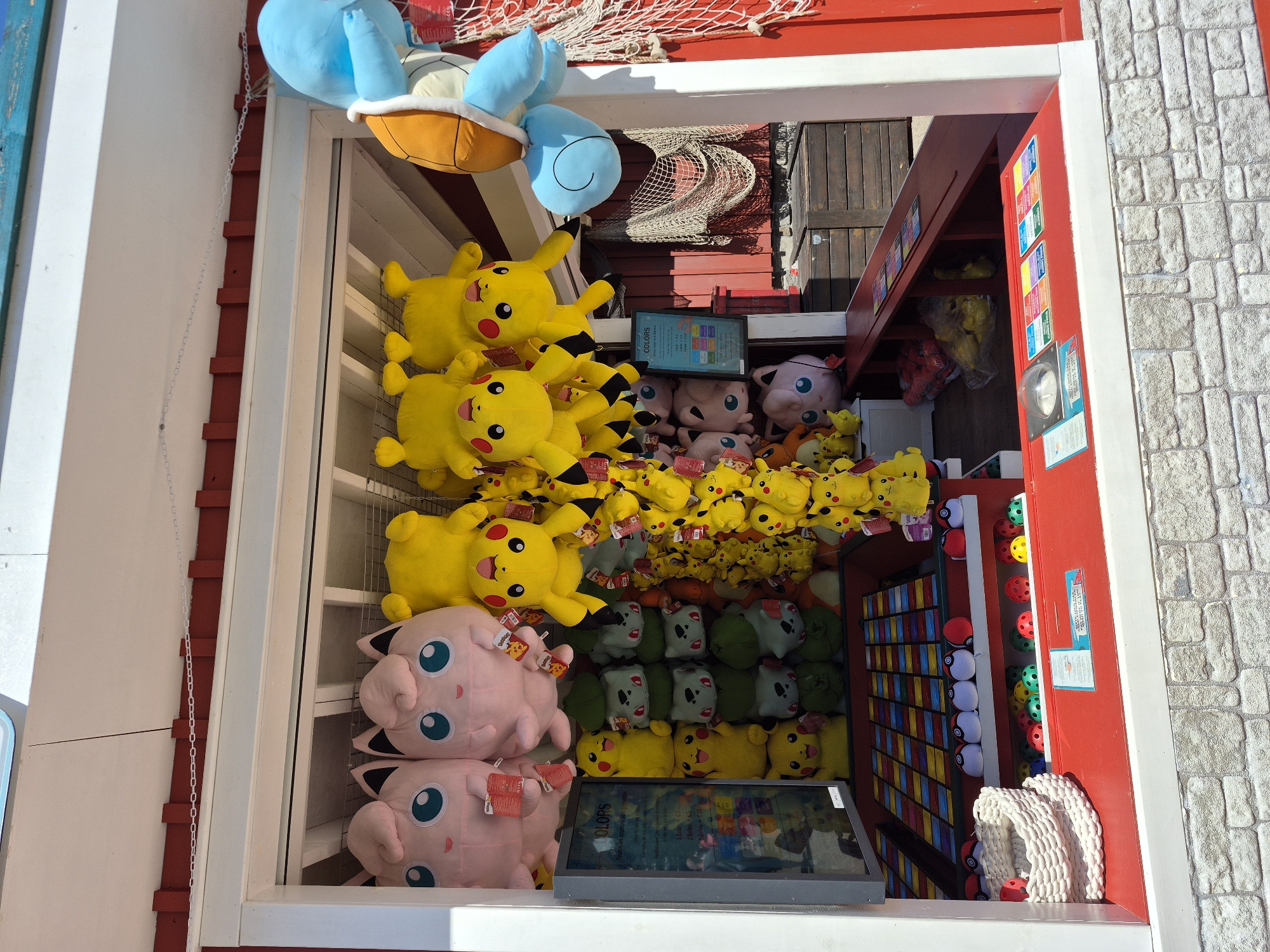 This image depicts a carnival game stall featuring various plush Pokémon toys and game elements. The stall has a red exterior with white trim and a white roof. Inside, numerous yellow Pikachu plush toys and pink Jigglypuff plush toys are hanging from the ceiling. On shelves below, more Pikachu, Jigglypuff, and green Bulbasaur plush toys are displayed. A blue Squirtle plush toy is visible hanging on the right side. A sign with the word "COLORS" is mounted on the wall. Various balls, some resembling Poké Balls, are arranged on shelves and in bins. The game stall is set up outdoors, with wooden crates visible in the background. The overall scene suggests a festive atmosphere at a fair or amusement park. This stall is located in Rust, Germany, a town known for the Europa-Park, a popular theme park where such attractions are common.