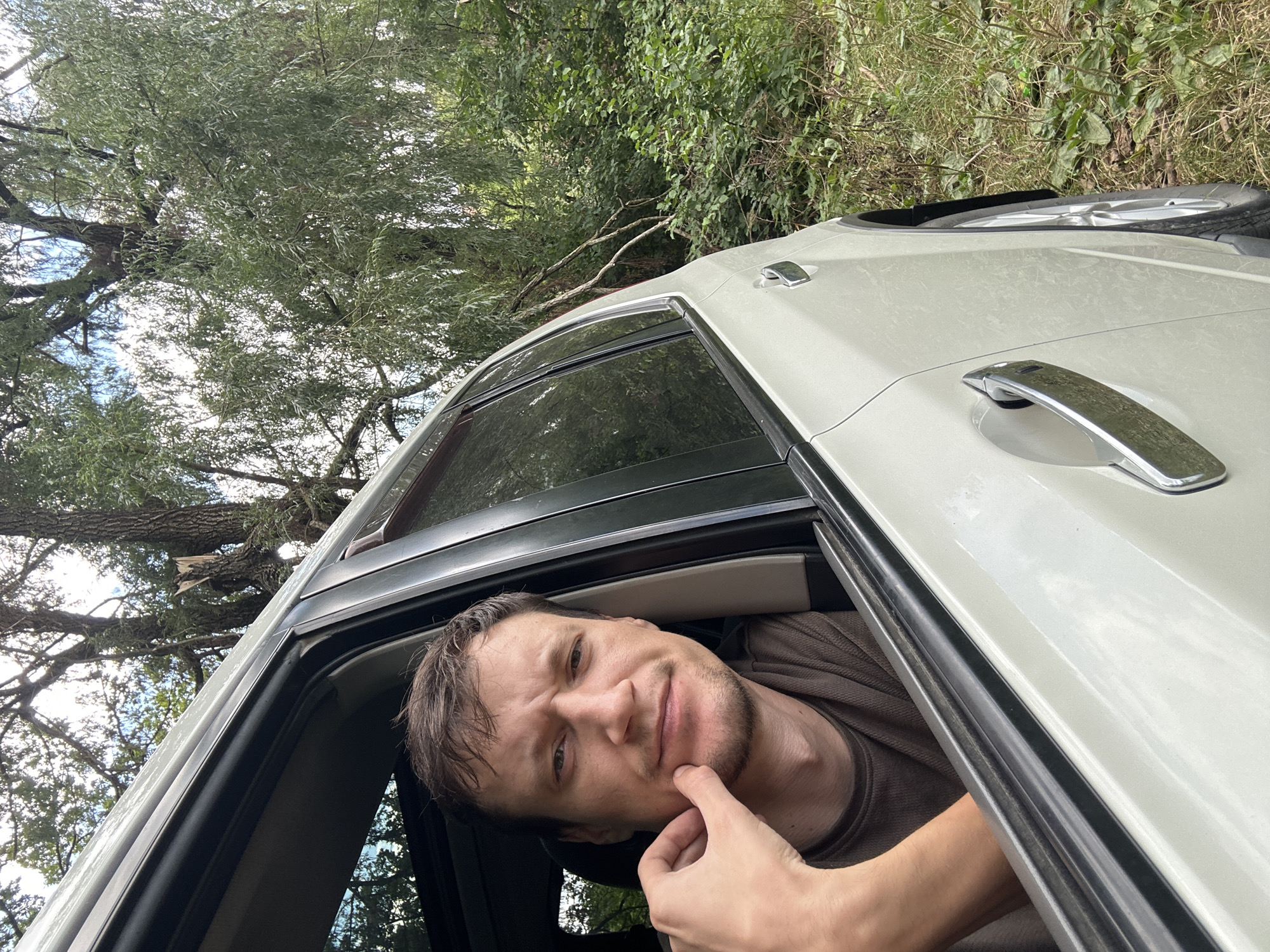 A man is looking out the driver's side window of a white SUV parked on a dirt road or shoulder. He is resting his chin on his hand and looking at the camera with a slightly furrowed brow. The car's interior is dark, and the man is wearing a brown t-shirt. Outside the car, lush green trees and foliage are visible, suggesting a natural or rural environment. There are no visible buildings or city structures, making it unlikely to be within the city of Makiv, Ukraine, unless it's on the outskirts or in a park area. The lighting suggests it is daytime.