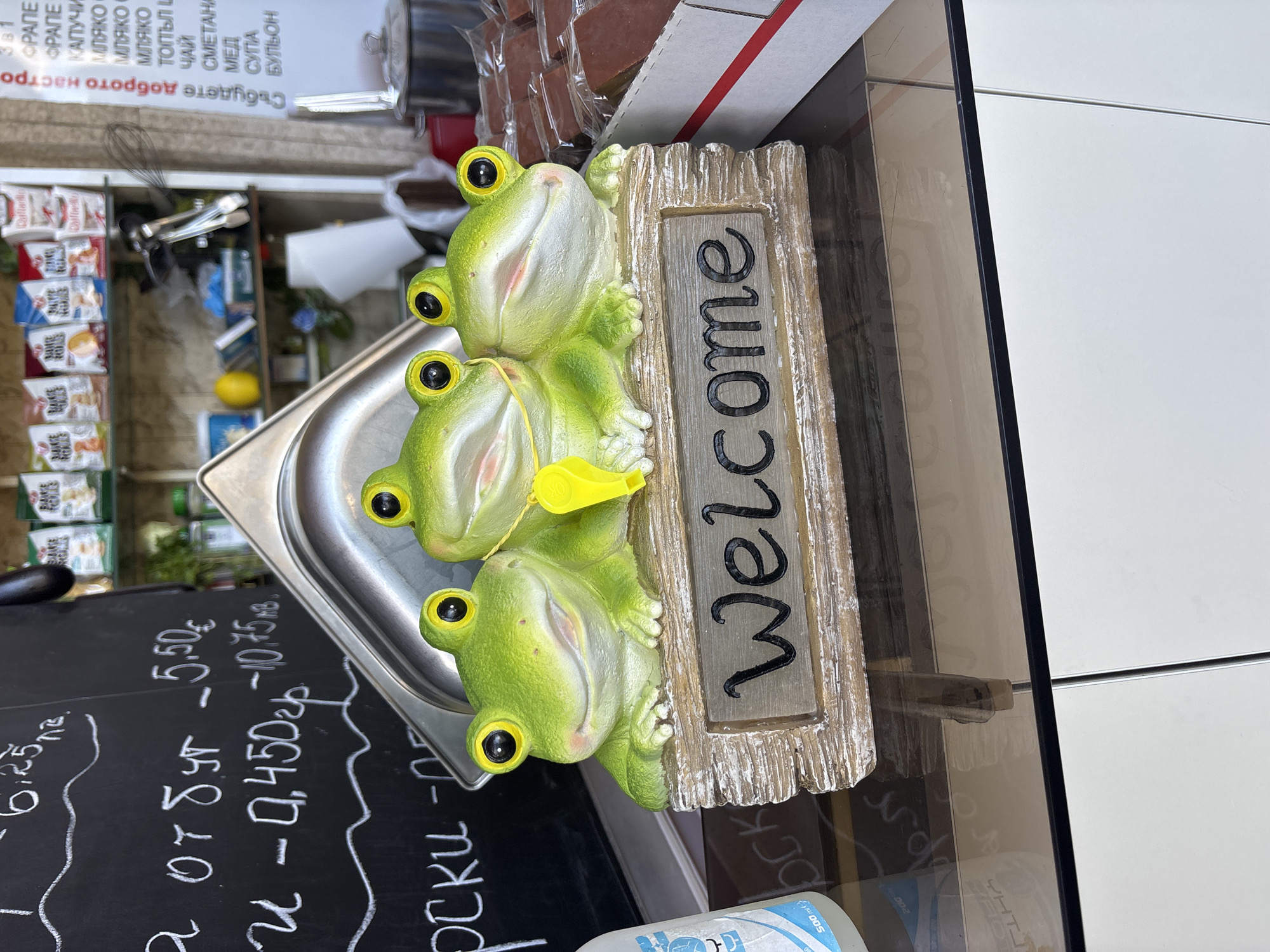 The image shows a close-up view of a decorative welcome sign featuring three cartoonish green frogs sitting on a wooden base. The sign is positioned on a reflective surface, likely a countertop, with a blurred background of shelves containing packaged food items and various kitchen utensils. The overall scene suggests a casual food establishment. The welcome sign is the primary subject, with the frogs having large, round, yellow-rimmed eyes and wide smiles. One frog has a yellow whistle hanging from its neck. The wooden base of the sign is weathered and has the word "Welcome" inscribed in black, stylized letters.
In the background, to the left, a portion of a chalkboard is visible, with chalk markings that appear to be prices and product names in Cyrillic. To the right, shelves are stocked with small bags of baked goods and other packaged food items. A metal container is visible in the center, and further to the right, a box of individually wrapped dark-colored food items, possibly chocolate bars or pastries, is partially visible.
The reflection on the countertop shows distorted shapes and colors from the surrounding environment, including what appear to be more chalk markings from the chalkboard. The lighting is bright, casting a slight glare on the reflective surfaces.
The specific location in the city of Sofia, Bulgaria cannot be determined from the visual information alone. The presence of Cyrillic script on the chalkboard and product packaging is consistent with Bulgaria, but does not pinpoint a precise location within Sofia.