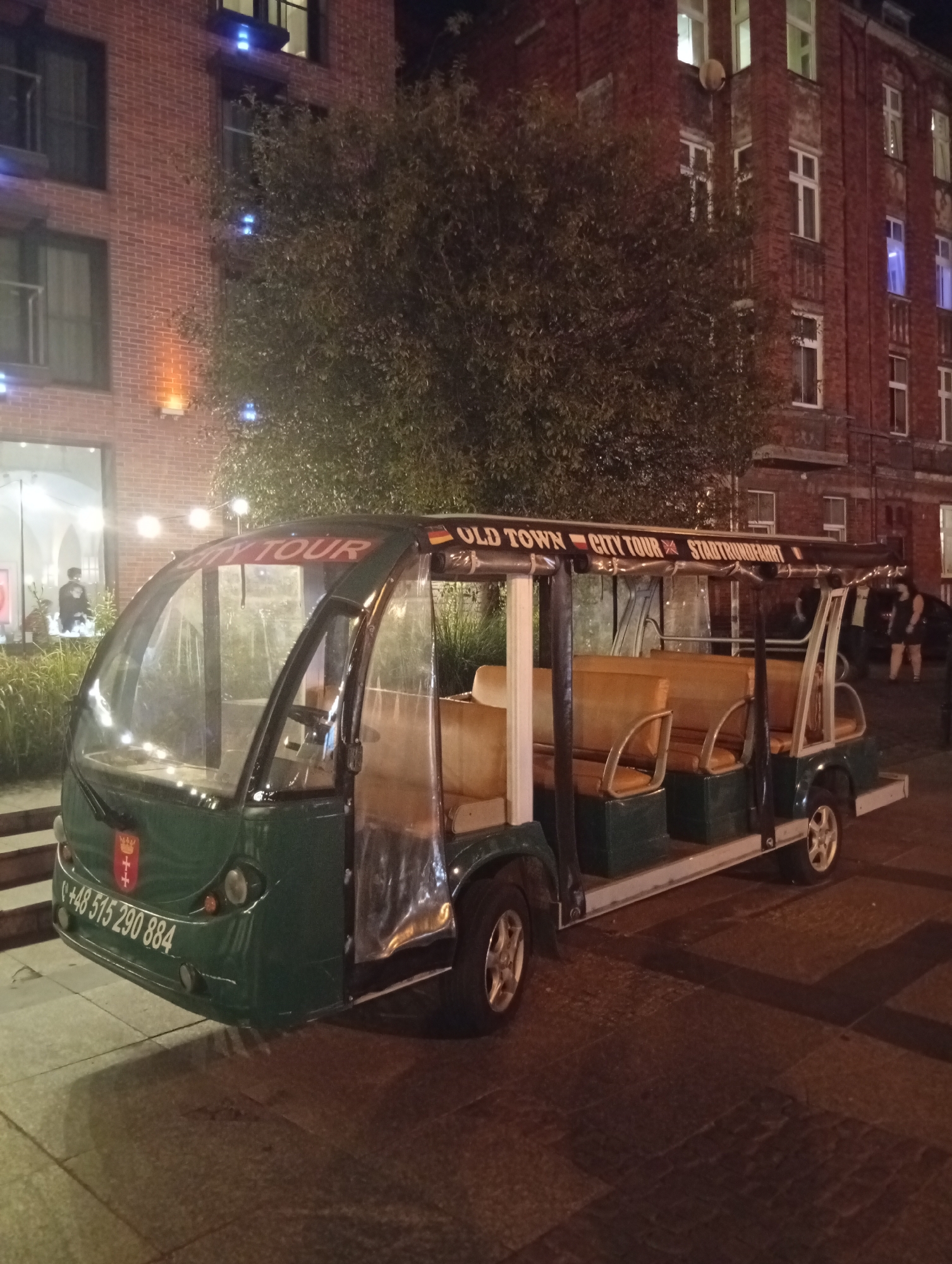 An open-air electric tour bus, dark green with beige seating, is parked on a paved sidewalk at night. The bus has a clear plastic enclosure around the front and sides, and a banner above the windshield reads "OLD TOWN CITY TOUR STADT RUNDFAHRT". A phone number, "+48 515 290 884", and a crest are visible on the front of the bus. The background features a brick building with several windows, some illuminated. A tree with dense foliage is situated between the bus and the building. In the distance, there are a few blurred figures of people and a parked car. The lighting suggests an urban environment at night. The crest on the bus is the coat of arms of Gdańsk, Poland, indicating the location is likely within the city of Gdańsk.