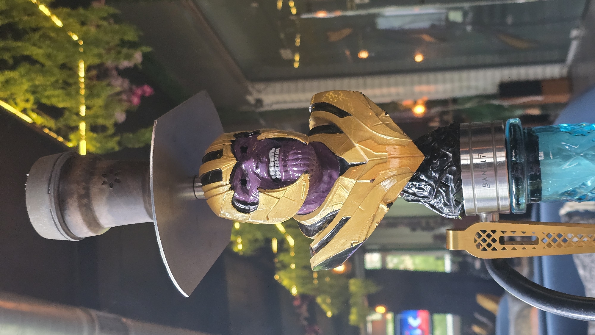 A close-up, outdoor, eye-level shot depicts a Thanos-themed hookah on a reflective surface in Sofia, Bulgaria. The hookah features a custom head with Thanos' head and gauntlet, constructed with gold and purple details. It is attached to a cylindrical metal body, a metal base plate, a black hose attachment, and a blue glass water chamber. The background reveals blurred cityscape reflections on the surface, including building lights, trees with string lights, and a pink flowering tree, suggesting an urban setting in Sofia during the evening.