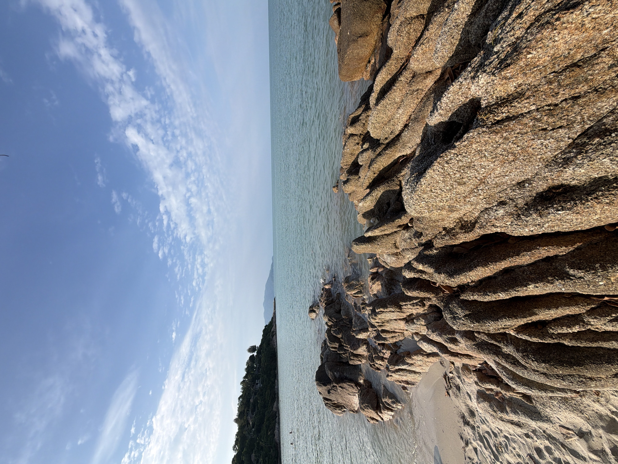 The image captures a scenic view of a rocky coastline in the Koh Samui District, Thailand. In the foreground, large, weathered rocks with a rough texture are partially submerged in the clear, shallow water. The sand on the beach is visible to the left of the rocks, showing ripples from the receding tide.
The water extends to the horizon, meeting a pale blue sky dotted with wispy white clouds. In the mid-ground, a lush, green hillside is covered with trees and some structures, suggesting a resort or residential area. Further in the distance, a mountainous landscape is visible under the hazy sky.
Two individuals are faintly visible in the water on the left side of the image, appearing to be swimming or wading. The overall scene is serene and natural, showcasing the coastal beauty of the region. The lighting suggests it is either early morning or late afternoon, with the sun casting soft shadows.