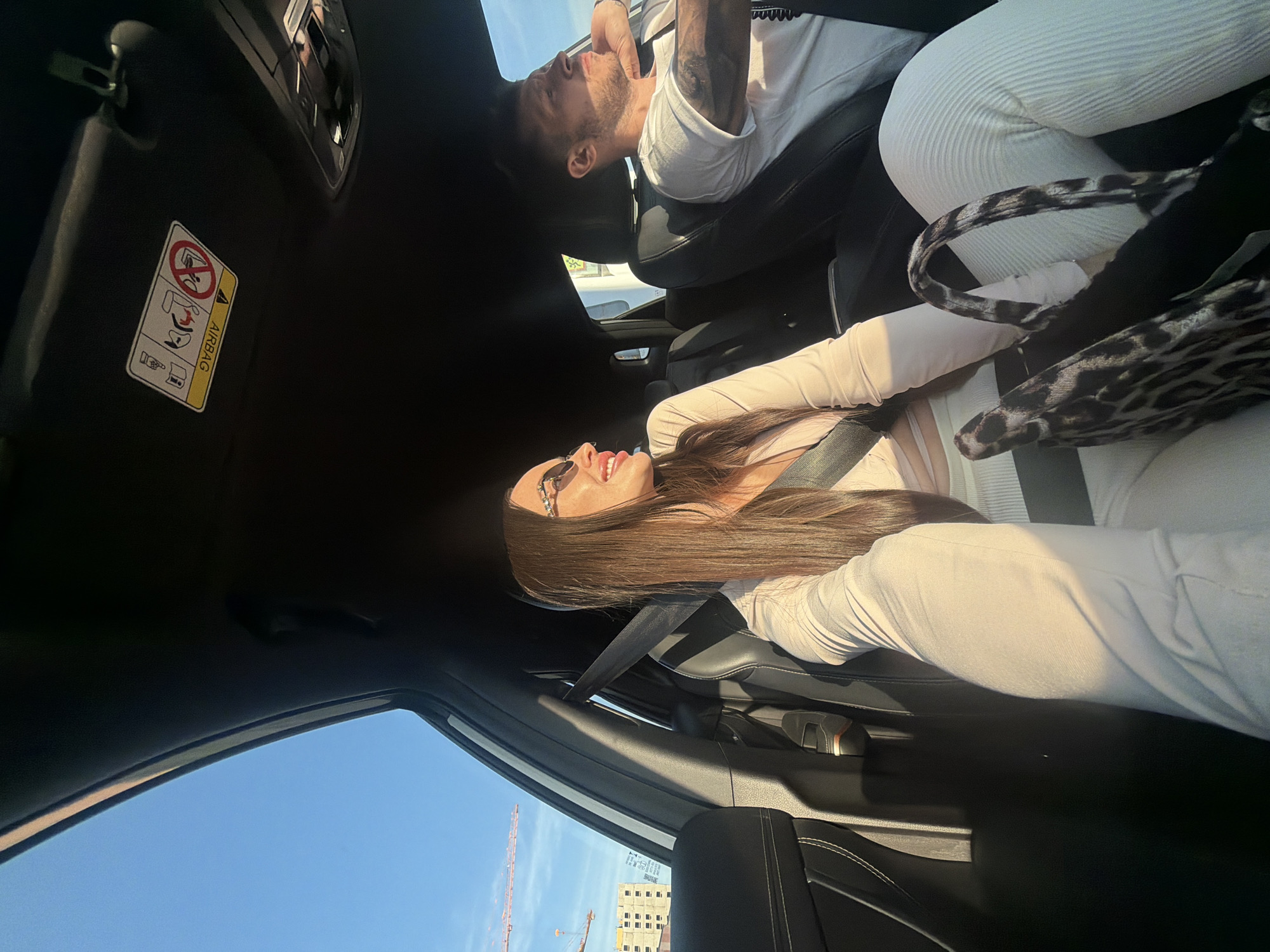 The image depicts the interior of a dark-colored vehicle, likely a car, during daytime with bright sunlight, situated in the city of Unknown, Russia. A woman with long brown hair, wearing sunglasses with embellished frames and a light-colored long-sleeved top and matching ribbed pants, is seated in the front passenger seat. She is secured by a seatbelt across her chest and is smiling while looking towards her right, out of the window. A leopard print handbag rests on her lap. In the driver's seat, a man with short dark hair and a beard, wearing a white t-shirt, is visible. He has tattoos on his left arm and is looking forward, with his right arm resting on the car's door frame. Through the windows on the passenger side, a clear blue sky, several large construction cranes, and multi-story buildings under construction are visible, indicating an urban or suburban environment within the city. The car's interior features dark upholstery, a sun visor with an upside-down 'AIRBAG' warning label (displaying icons related to child seats and airbags), and an overhead light console. Strong sunlight casts distinct shadows within the vehicle cabin.