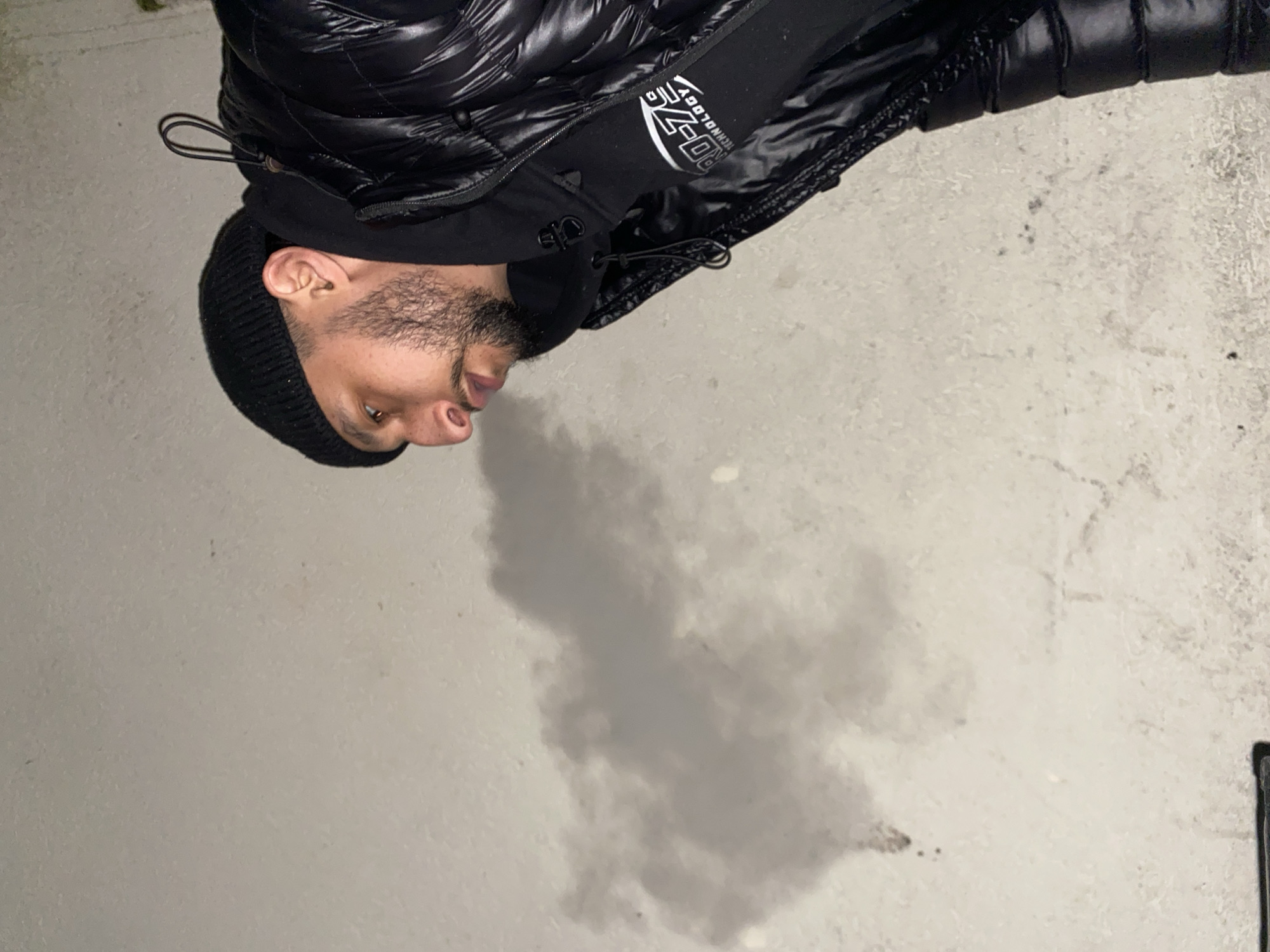 The image depicts a man wearing a black beanie, a black hooded sweatshirt with the text "RD-Z Technology", and a black puffer jacket. The background appears to be a concrete surface, possibly a sidewalk or wall. There is a dark, indistinct smudge or discoloration on the surface. The scene is likely an urban environment, but the location cannot be confirmed as Schiltigheim, France based on the visual information.
