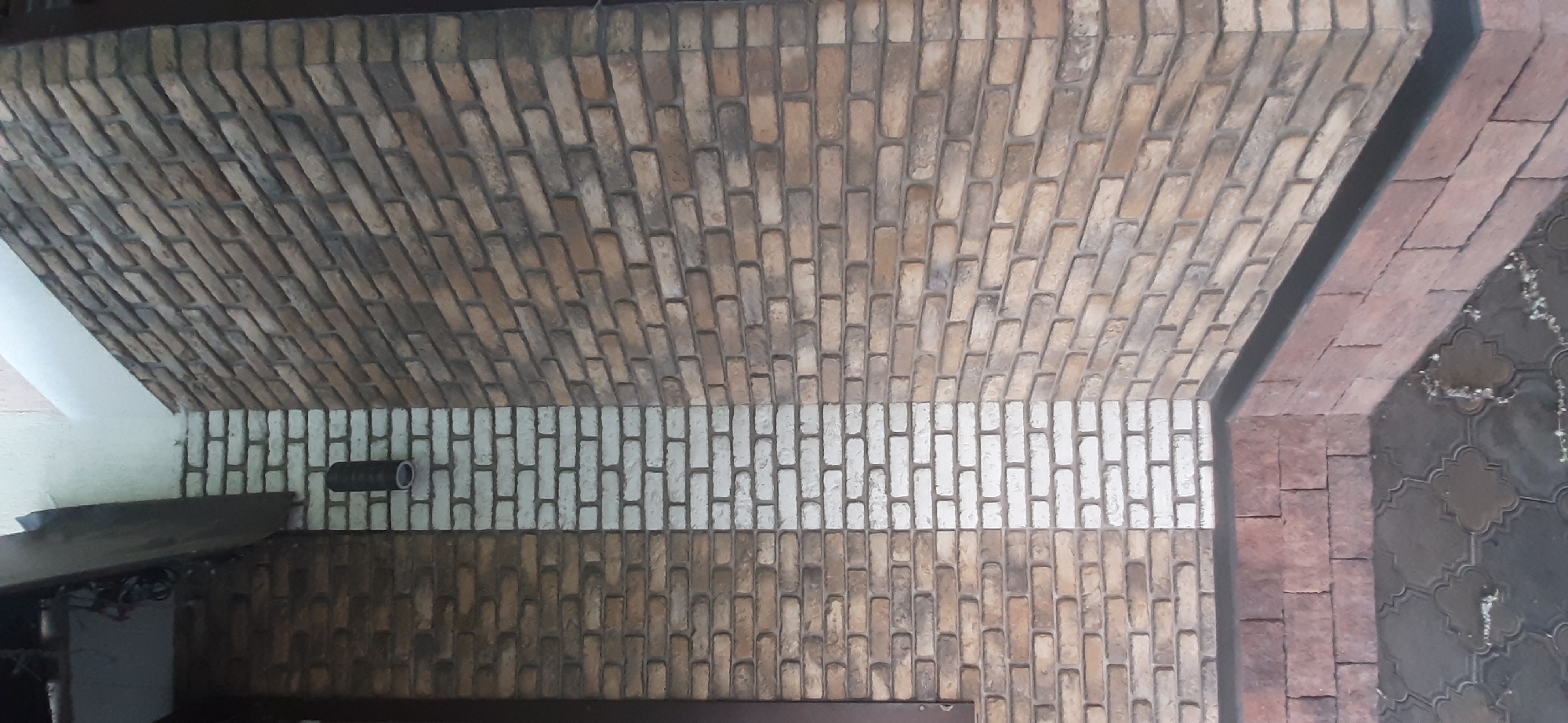 The image shows a close-up view of the exterior corner of a building. The walls are constructed of brick, with a vertical stripe of white brick separating sections of brown and tan brick. A dark cylindrical light fixture is mounted on the white brick stripe. The lower portion of the walls, near the ground, is faced with reddish-brown brick. The ground in front of the building is paved with dark grey interlocking pavers with a decorative pattern. The scene appears to be outdoors. This image was taken in Yessentuki, Russia.