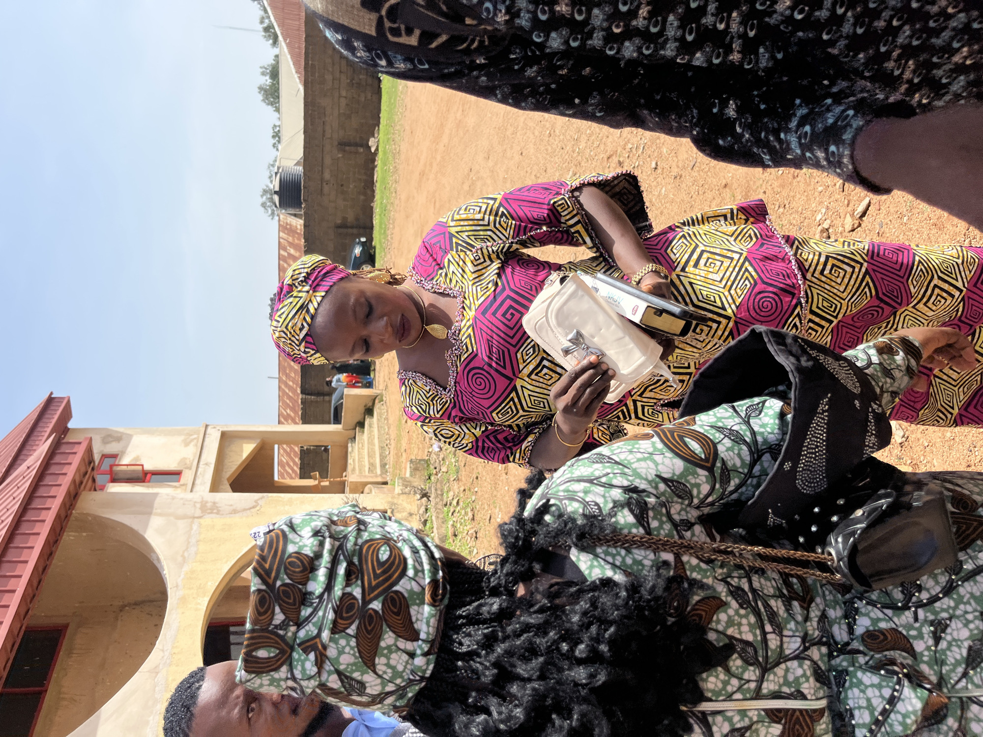 In Bukuru, Nigeria, three individuals are visible outdoors, standing in front of a light-colored, multi-story building with a red roof. The ground is covered in reddish-brown dirt and gravel.
On the left, a man is partially visible, looking towards the right. His left shoulder and the side of his head are in frame. He is wearing a light blue shirt.
In the center, a woman is facing slightly to the left, looking down at items in her hands. She is wearing a colorful, patterned outfit consisting of a headscarf and a dress with a square and circular geometric design in pink, yellow, and black. She is holding a cream-colored clutch bag and what appears to be a book or papers. She wears gold earrings and a gold necklace. A thin gold bracelet is visible on her right wrist.
On the far right, a person is mostly obscured, with only their shoulder and arm visible. They are wearing a dark, patterned fabric.
To the left of the central woman and in front of the man, another individual is seen from behind, wearing a green and white patterned headscarf and a matching green and white patterned outfit with brown and black accents. They have dark, curly hair visible. This person is holding a black purse with a chain strap, which is adorned with rhinestones and studs.