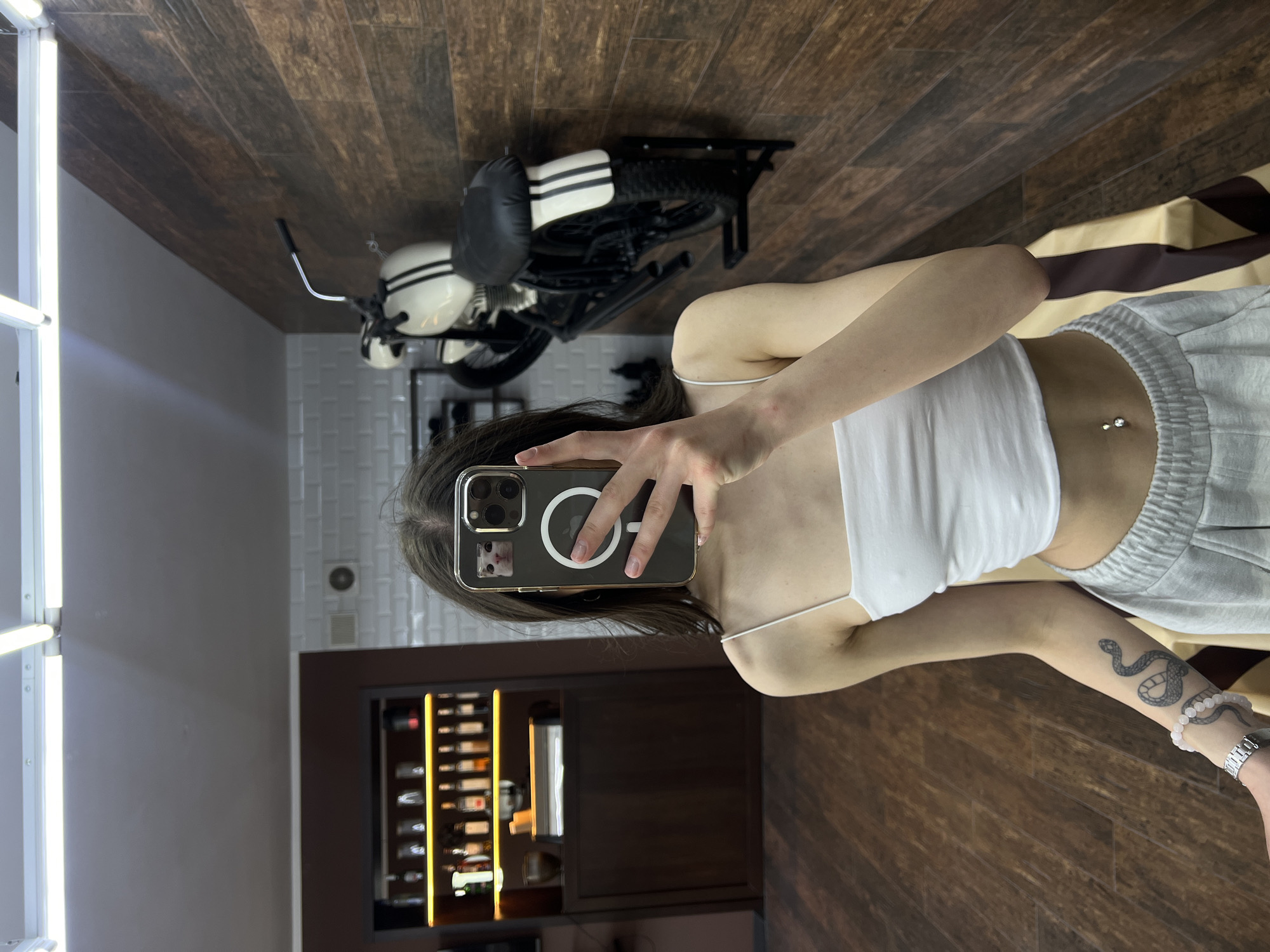A person is taking a selfie indoors. The individual is wearing a white tank top and gray sweatpants. The phone case has a magnetic charging circle and a picture of a cat. A snake tattoo is visible on the person's arm, along with a bracelet and a watch. In the background, there's a motorcycle mounted on the wall, a wine rack, and a white brick wall with a light fixture. The background also reveals wooden flooring. While the city of Ternopil, Ukraine, cannot be verified directly from the image, there is no information in the image to contradict it being taken there.