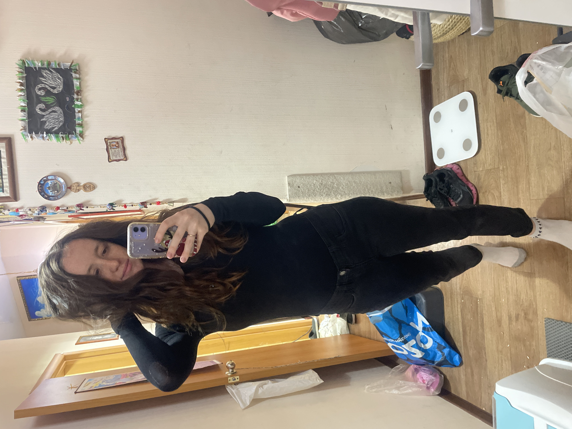 A young woman is captured taking a mirror selfie in an indoor setting, likely a bedroom or hallway. She is wearing a long-sleeved black top, black jeans, and white socks. Her long, dark brown hair cascades over her shoulders, partially obscuring her face as she holds a smartphone with a patterned case to capture her reflection. A ring is visible on her right-hand ring finger.
The room features a light-colored wall decorated with various items, including a framed picture of two white swans on a dark background with a green and white fringe border, a smaller framed image, a decorative plate, and a carved wooden spoon. Below the mirror, a wooden shelf is visible, upon which a small metal fixture and some plastic bags rest.
The wooden floor shows a white digital bathroom scale, several pairs of sneakers (including black, green, and black with pink details), and multiple plastic bags. A prominent blue shopping bag has the text "YOYÓ" visible on its side. The overall scene suggests a personal and lived-in space, illuminated by what appears to be natural daytime light.
