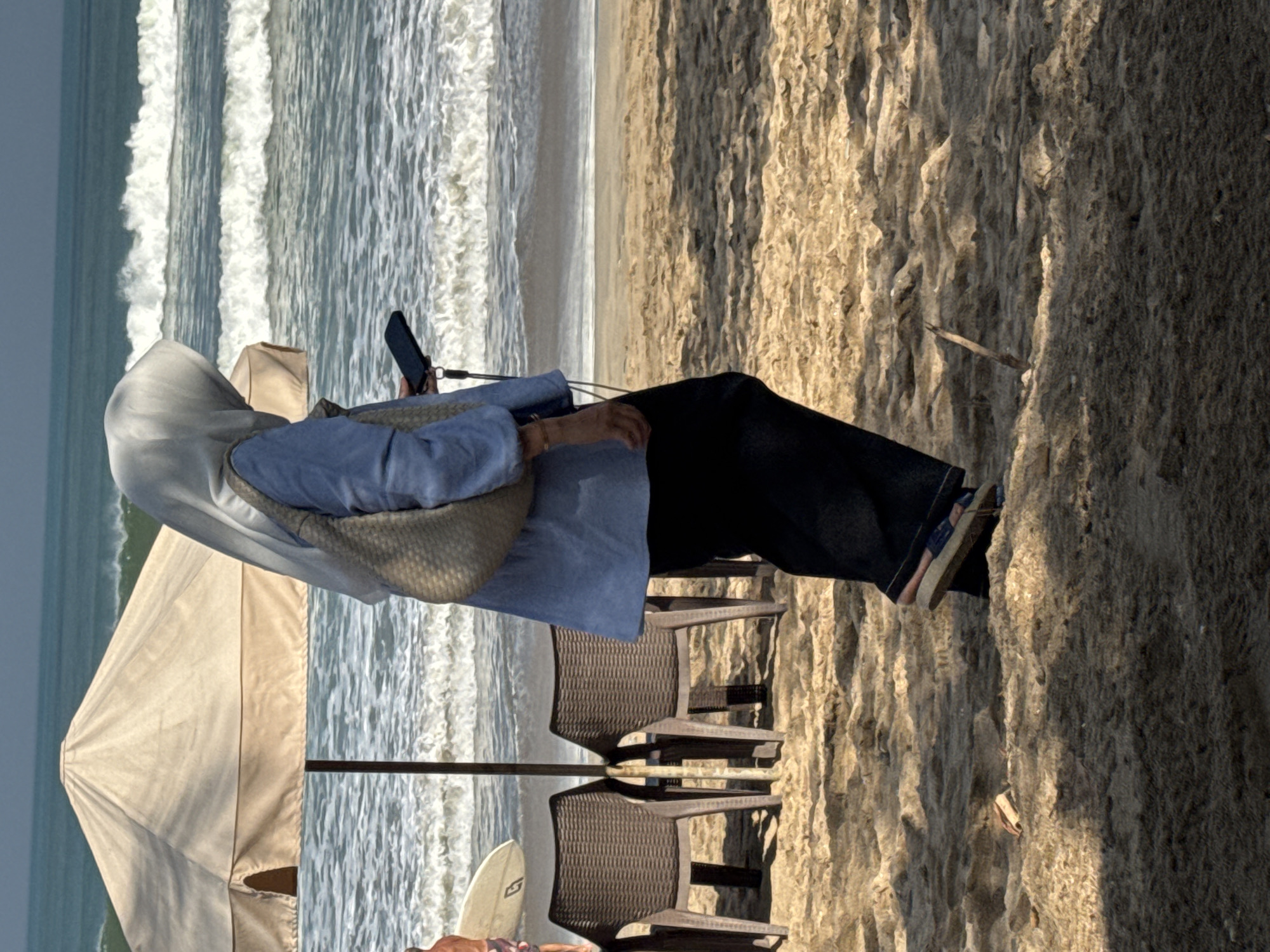 In Kuta, Indonesia, a person wearing a light blue long-sleeved shirt, black pants, and a light grey hijab walks on a sandy beach. The person is carrying a light brown woven bag over their shoulder and holding a dark-colored mobile phone in their right hand. A beige beach umbrella stands behind the person, with two empty brown plastic chairs positioned beneath it. The ocean, with visible waves, is in the background, and a white surfboard stands near the chairs. The sand is textured and shows shadows from the umbrella and surrounding objects.