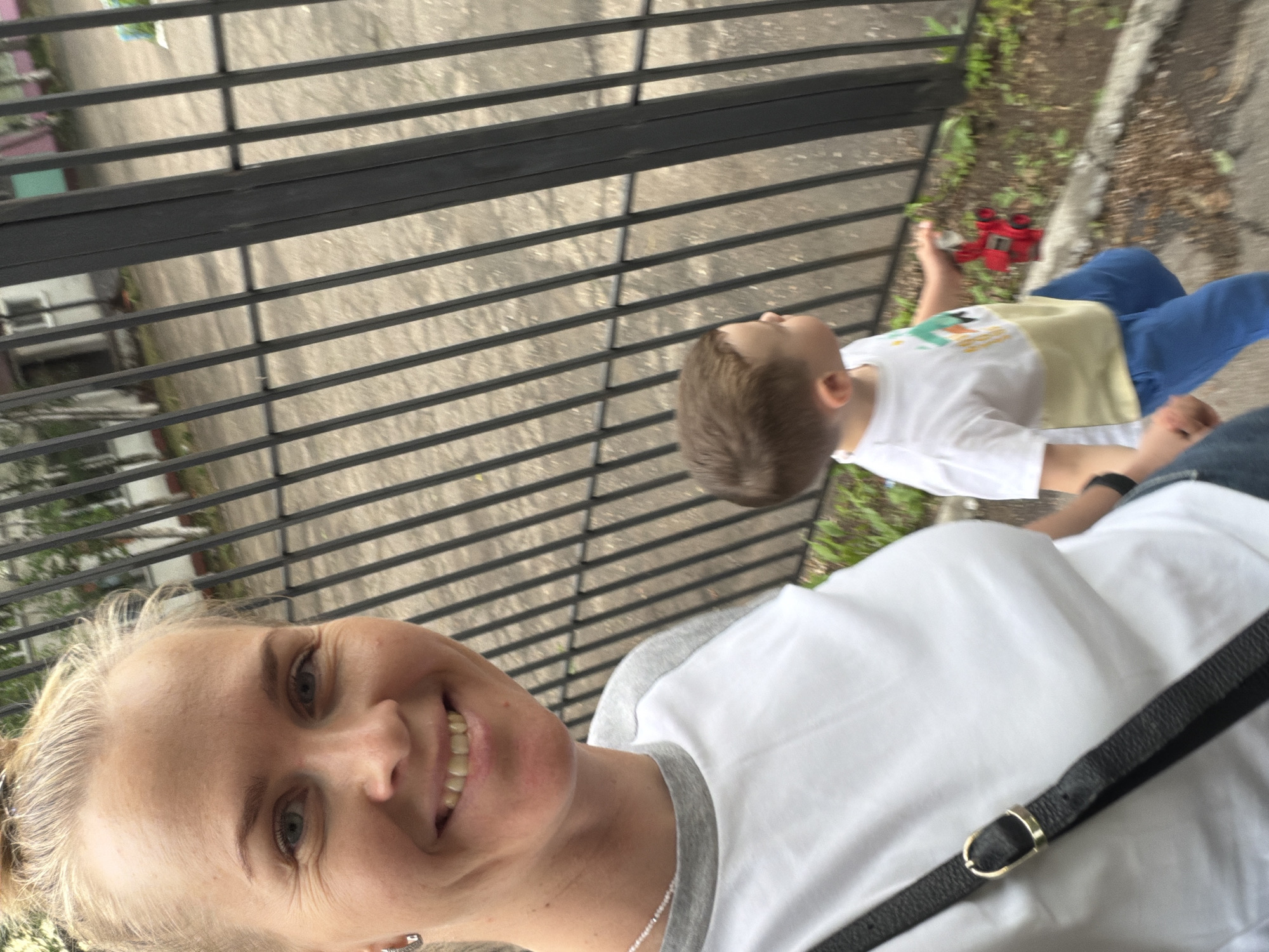 A woman and a young boy are captured in a selfie taken from a low angle, outdoors. The woman, positioned in the foreground on the left, is smiling and looking towards the camera. She has blonde hair styled in a bun and is wearing a white t-shirt with grey sleeves and a grey neckline. A black crossbody bag with a gold buckle is slung across her shoulder.
To the right and slightly behind her, the young boy is walking away from the camera, looking to his right. He has light brown hair and is wearing a white t-shirt with a yellow and green design on the front, paired with blue trousers. He is holding a small red toy in his right hand.
They are walking along a paved path next to a black metal fence. Through the fence, a building with light-colored walls and windows is visible, along with some greenery and trees. The overall scene suggests a casual outdoor outing. The presence of a building with a pink section and the style of the fence are consistent with a common type of educational institution or public park found in Khabarovsk, Russia, potentially a kindergarten or schoolyard.