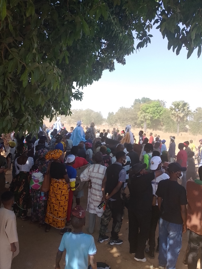 This image depicts a large outdoor gathering in Kpak, Nigeria, under a clear, bright sky. A dense crowd of individuals, including adults and children, occupies the foreground and midground, standing on dry, dusty ground. The upper left of the scene is shaded by the full green foliage of a large tree. In the midground, several individuals, notably three, are elevated above the main crowd, adorned in light-colored, flowing robes—one in light blue, two in cream or yellow—with distinct head coverings, suggesting a ceremonial or significant presence.
The crowd consists of diverse individuals dressed in a mix of traditional African attire, such as patterned dresses and headwraps on women, and modern clothing like t-shirts, trousers, and vests on men. Some individuals in the foreground are wearing face masks. One man in the foreground is dressed in a white and brown striped tunic, while another wears camouflage trousers and a dark vest, holding a dark bottle. In the immediate foreground, a young boy in a light blue t-shirt stands with his back to the camera. The background features sparse vegetation, including distant trees and what appears to be a palm tree, under a slightly hazy sky, consistent with a semi-arid or savanna environment. The people are largely static, observing the elevated figures in the midground, indicating a communal event or procession.