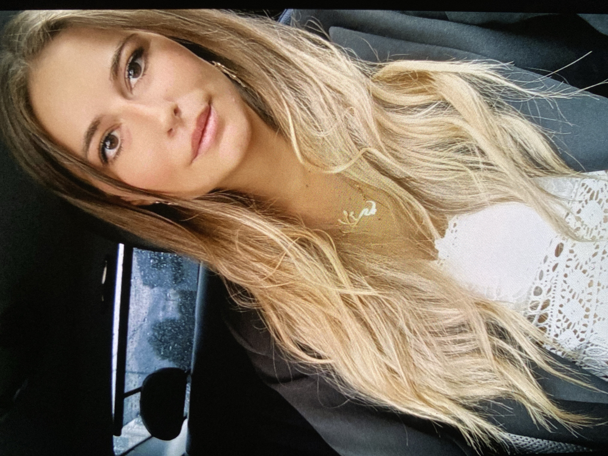 A head-and-shoulder portrait features a young woman with long, flowing blonde hair, cascading over her shoulders and chest. Her hair is multi-toned with natural highlights and appears slightly tousled. She has fair skin, dark brown eyes, and light brown eyebrows, accentuated by eyeliner and a soft pink lip color. Her expression is calm and direct, with a gentle, slight smile as she looks towards the viewer.
She is wearing a light-colored, possibly white or cream, top featuring a delicate, lacy, open-weave pattern visible on her right side. A thin, gold-toned necklace with a small, stylized script-like charm (resembling the word "love") is visible on her neck, and a gold earring is partially seen on her right ear.
The setting places her inside a vehicle, indicated by the dark, upholstered seats visible in the background. A car window on her left shows raindrops on the glass, suggesting it is raining or has recently rained, and the blurred outdoor view reveals dark, possibly wet, foliage. The overall lighting is dim, indicative of an overcast day. This casual portrait is captured in Bankya, Bulgaria.
