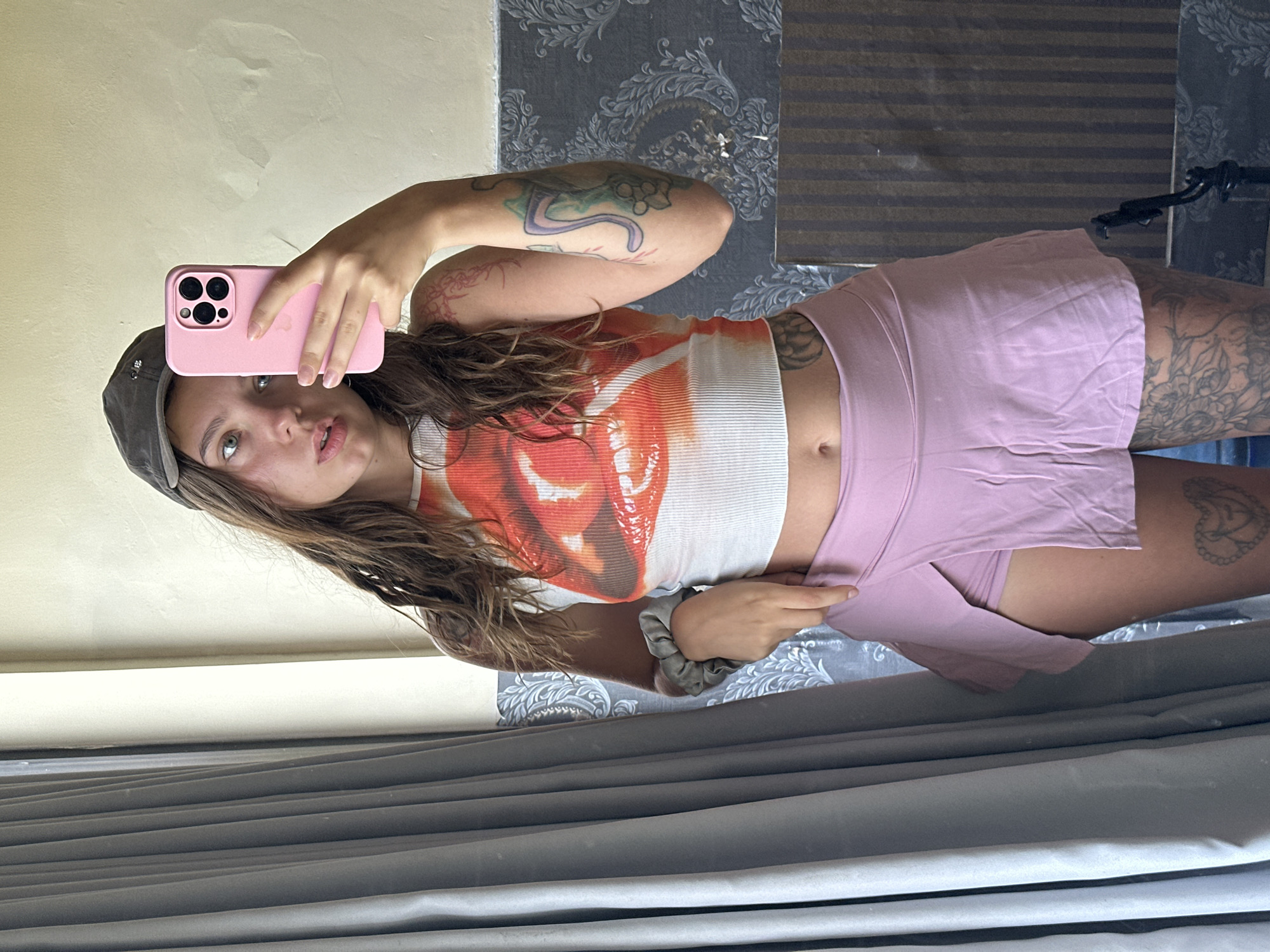 A young woman with long, wavy brown hair, wearing a dark baseball cap, a white cropped tank top with a red and orange lip print, and a light pink skort, is taking a selfie in a mirror. She is holding a pink iPhone in her right hand, which is raised to take the photo. Her left hand is near her waist, with her fingers slightly curled. She has tattoos on her arms and legs. The background shows a grey curtain on the left and a wall with textured wallpaper on the right. The location is indoors, with neutral-colored walls.