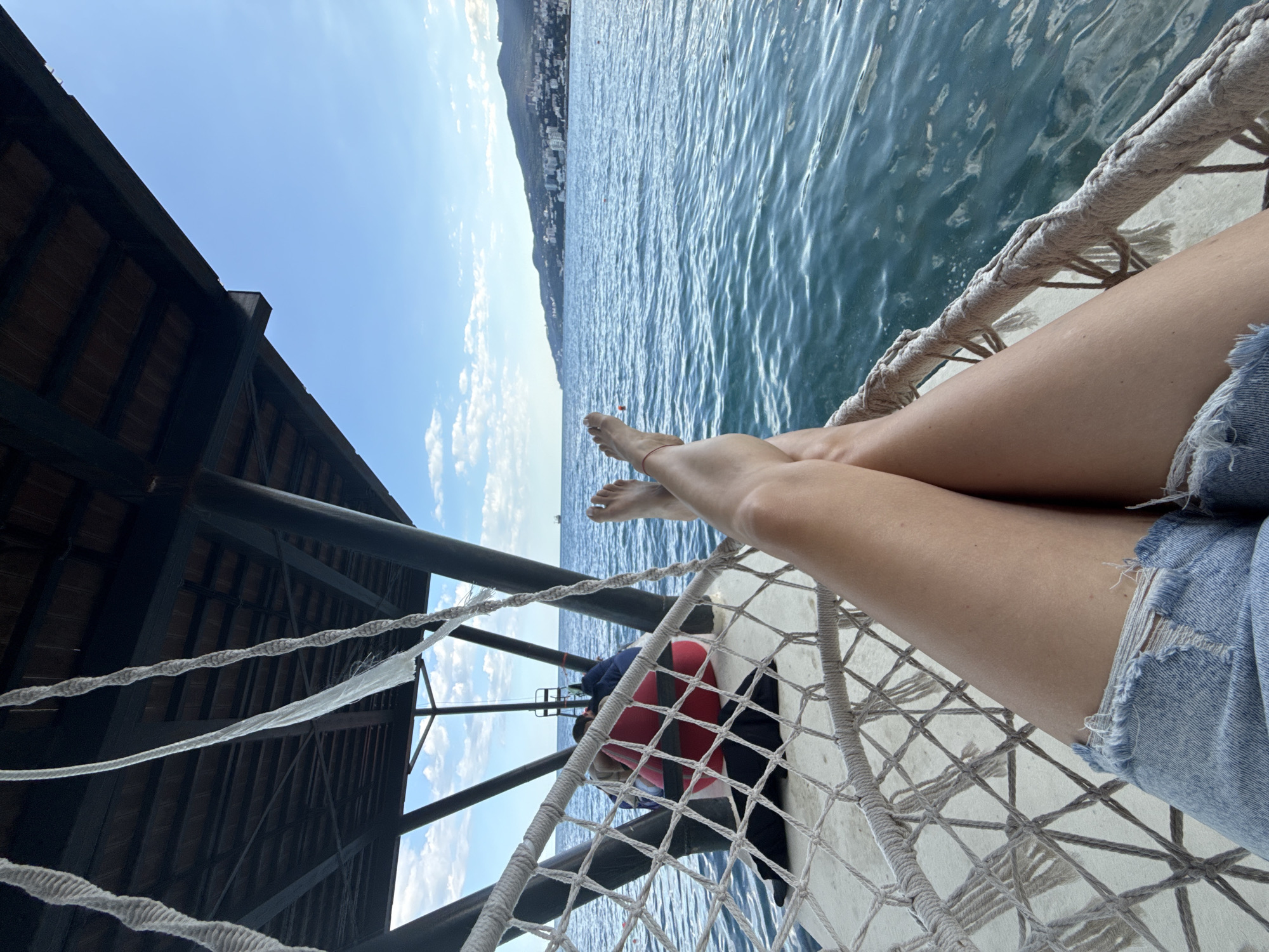 This is a first-person perspective media file, likely a selfie, taken on a sunny day with a clear blue sky and scattered clouds. The scene overlooks a calm, blue body of water, possibly a sea or large lake, with gentle waves. In the distance, a coastline with hills and buildings is visible, suggesting an urban or resort area. The air appears clear, and the overall mood is relaxed and serene.
The subject, whose legs and feet are prominently featured, is resting on what appears to be a hammock or a net-like structure suspended over the water. They are wearing denim shorts that are ripped at the hem. One foot has a thin red string bracelet on the ankle.
To the left of the frame is a dark wooden structure, possibly part of a pier, a balcony, or a covered seating area, with visible support beams and a slatted roof. Ropes from the hammock or netting crisscross the lower part of the image. A red and white life preserver is visible below the hammock.
The time of day is likely daytime, given the bright sunlight and blue sky. The location is identified as Yalta, Ukraine. The activity is passive enjoyment of the scenery, with the subject likely relaxing and taking in the view. No visible text is present in the media file.