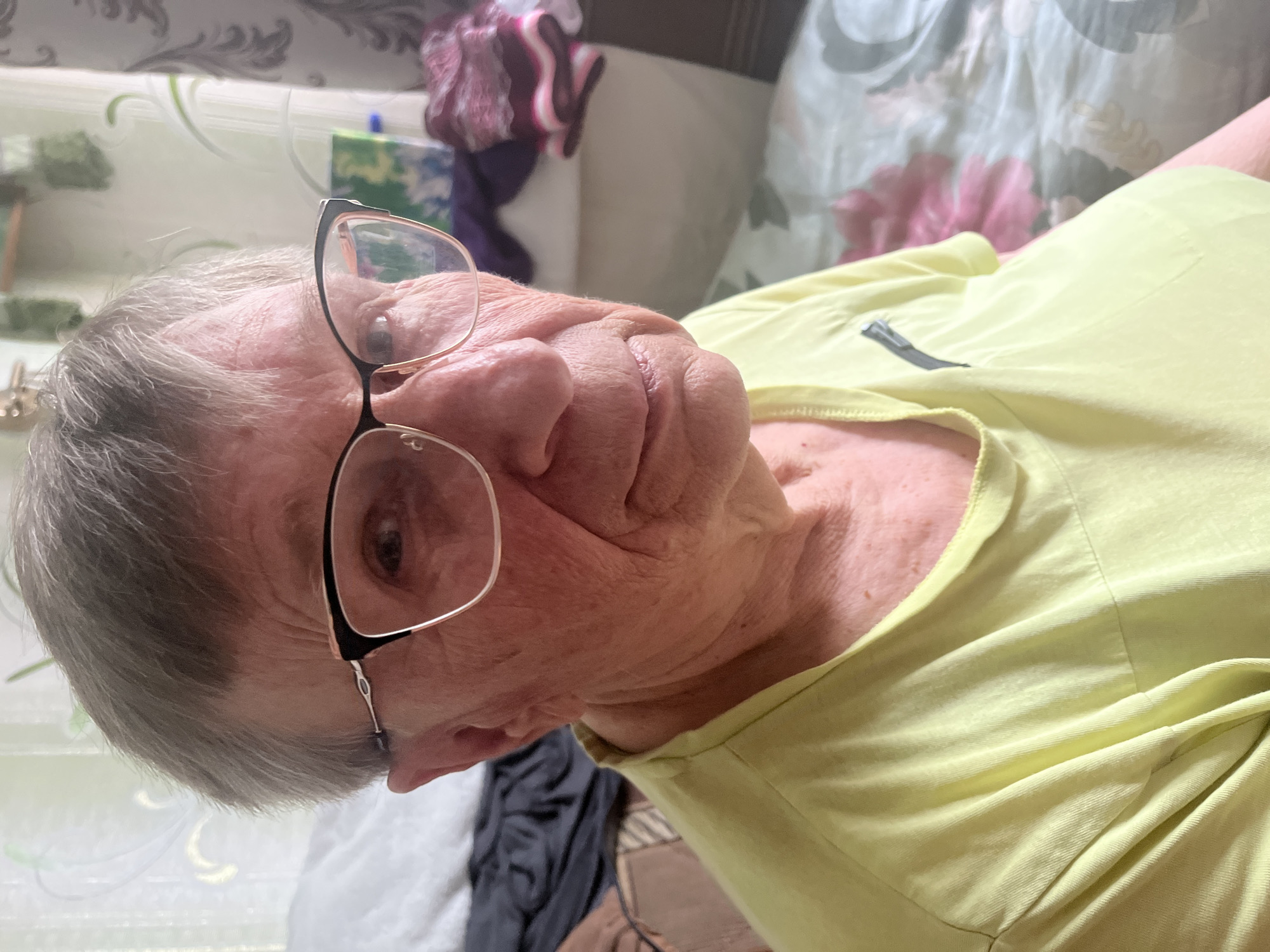 A close-up, slightly angled, medium-shot portrait of an elderly Caucasian woman wearing glasses and a light yellow t-shirt. The woman has short, gray hair and is looking towards the camera with a neutral expression. The background appears to be an indoor setting, possibly a bedroom, with a patterned comforter on a bed. The lighting is natural and diffused, suggesting it's daytime. The image lacks any clear indication of a specific location within Minsk, Belarus, or any particular action or interaction taking place.