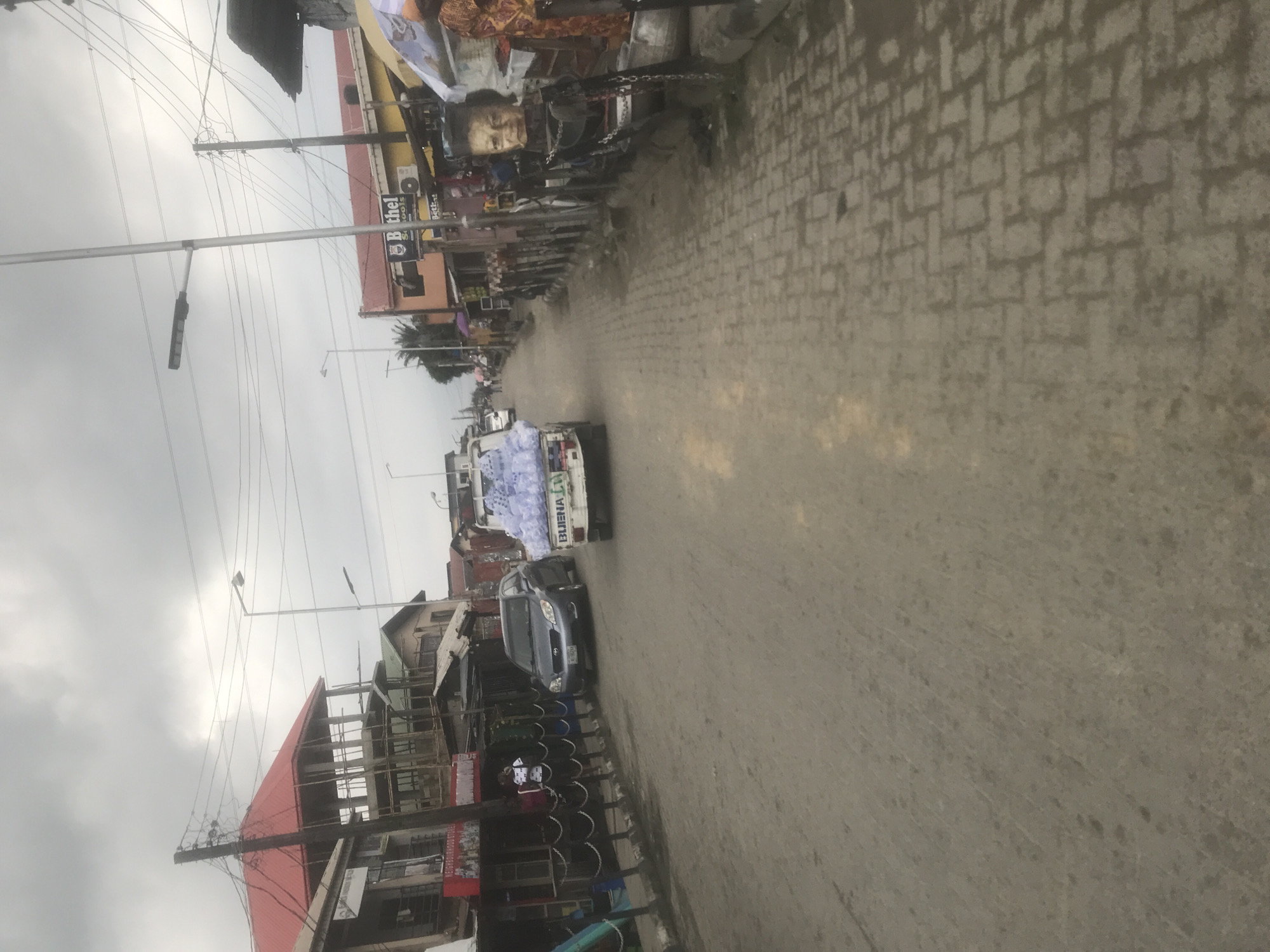 This media file captures a street scene, likely in an urban or semi-urban setting, with buildings lining both sides of a paved road. The image is oriented vertically.
On the left side of the road, a row of buildings is visible. Some appear to be shops, with open fronts displaying goods. One building has a prominent red roof and scaffolding, suggesting ongoing construction or renovation. Another building has a sign that partially reads "Bethel...ools". Overhead, a complex network of electrical wires and poles spans across the scene.
On the right side of the road, the pavement is made of interlocking bricks. A light-colored truck is partially visible, loaded with what appears to be white plastic sheeting or packaging. A dark-colored car is parked behind it. Further down the street, more buildings and shops are seen. There are some figures of people visible, but they are small and indistinct. One stall displays what looks like a mannequin or a sculpture of a human face with exaggerated features.
The weather appears overcast, with a dull sky. The lighting suggests it could be daytime, but the clouds limit bright sunlight. The overall atmosphere is that of a busy street, possibly a marketplace, with commercial activity and traffic. There is some visible text on signs and on the truck, but it is not clearly legible in most cases. One sign on the truck reads "BUENA LTO".